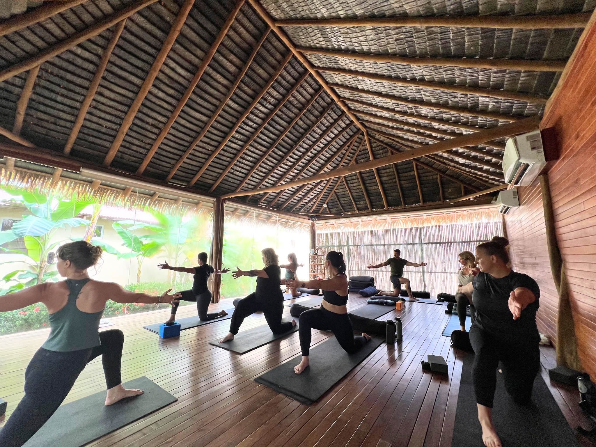 A group of people practicing yoga in a spacious, open-air room with a high, thatched roof and large open windows overlooking greenery. Participants are performing yoga poses on mats.