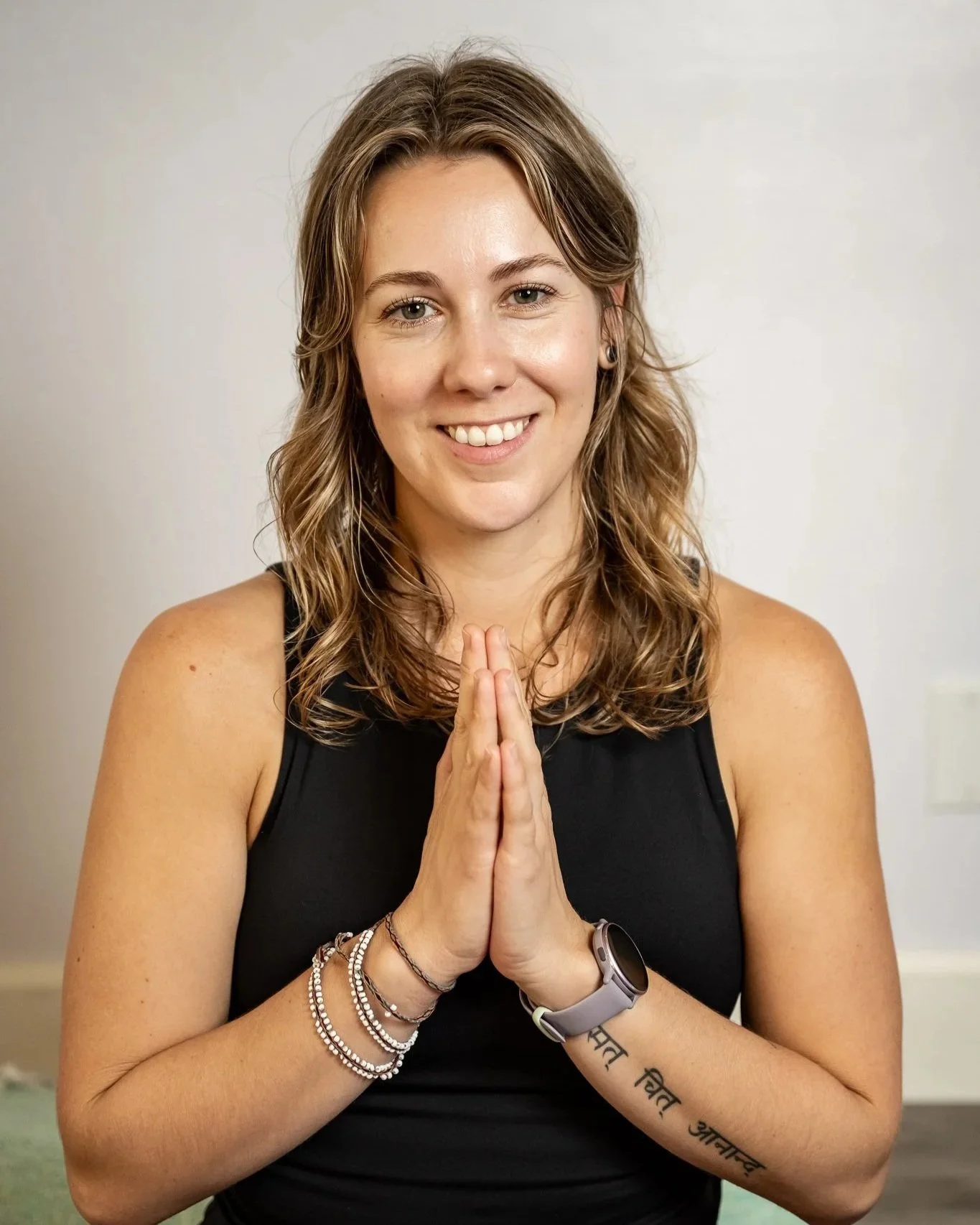 A woman with wavy brown hair, wearing a black tank top, standing indoors against a plain white wall. She is smiling, with hands pressed together in a prayer-like pose.