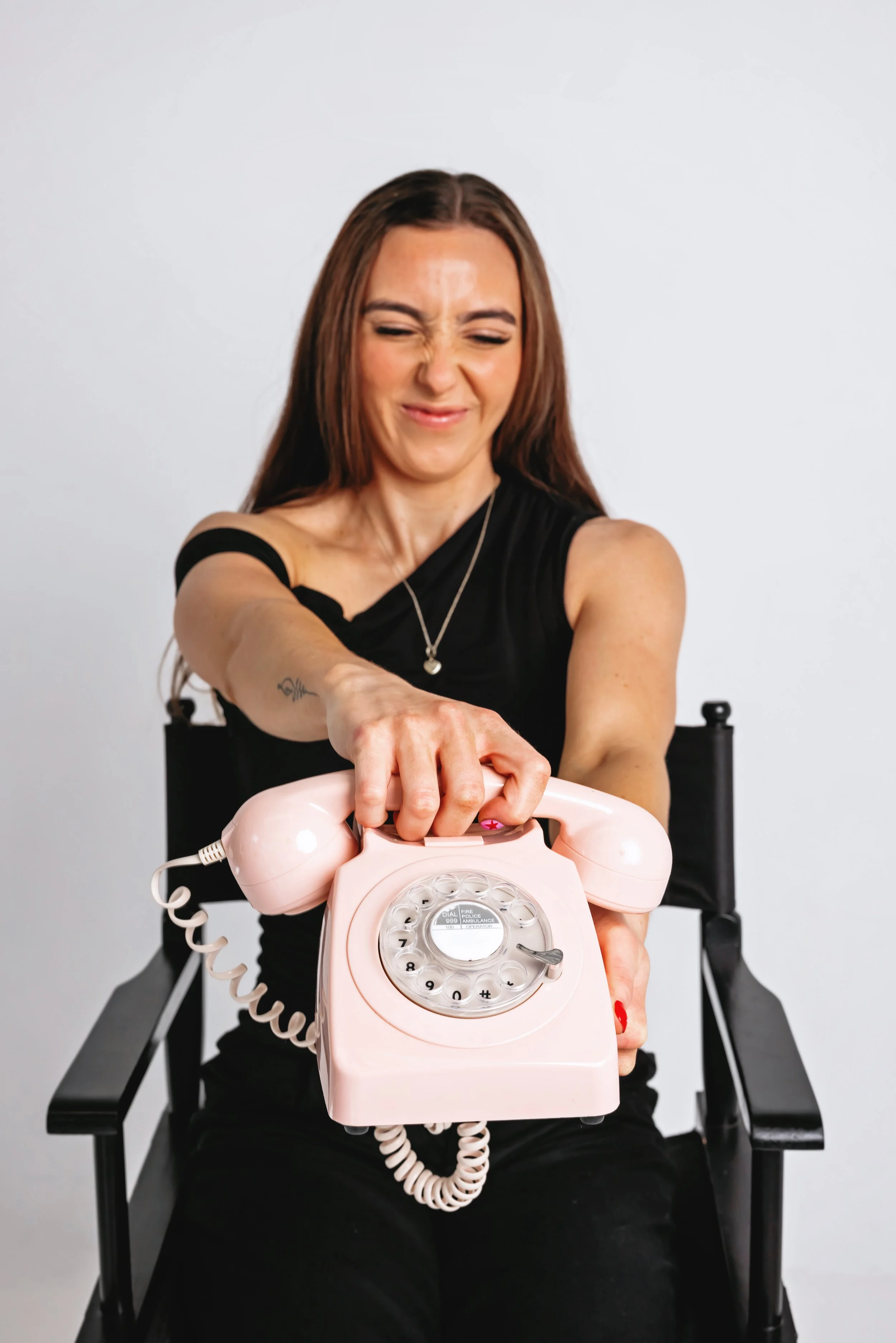 A woman with long brown hair smiling and grimacing while reaching to pick up a pink rotary phone, seated on a black director's chair against a plain white background.