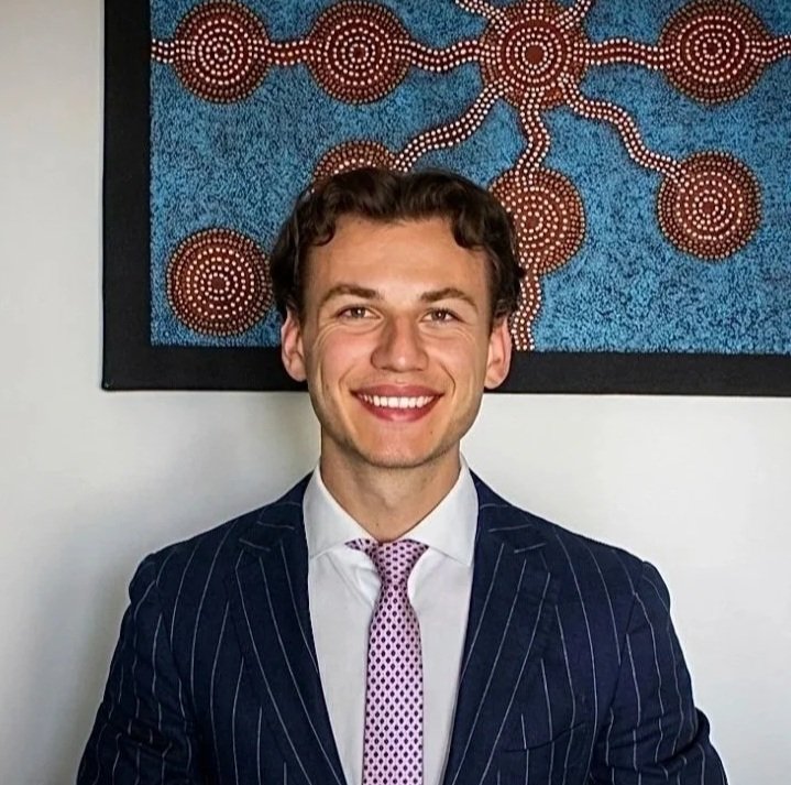Lewis Bradbury Young man in formal attire with pink shirt, orange tie, and dark blazer on a light gray background.