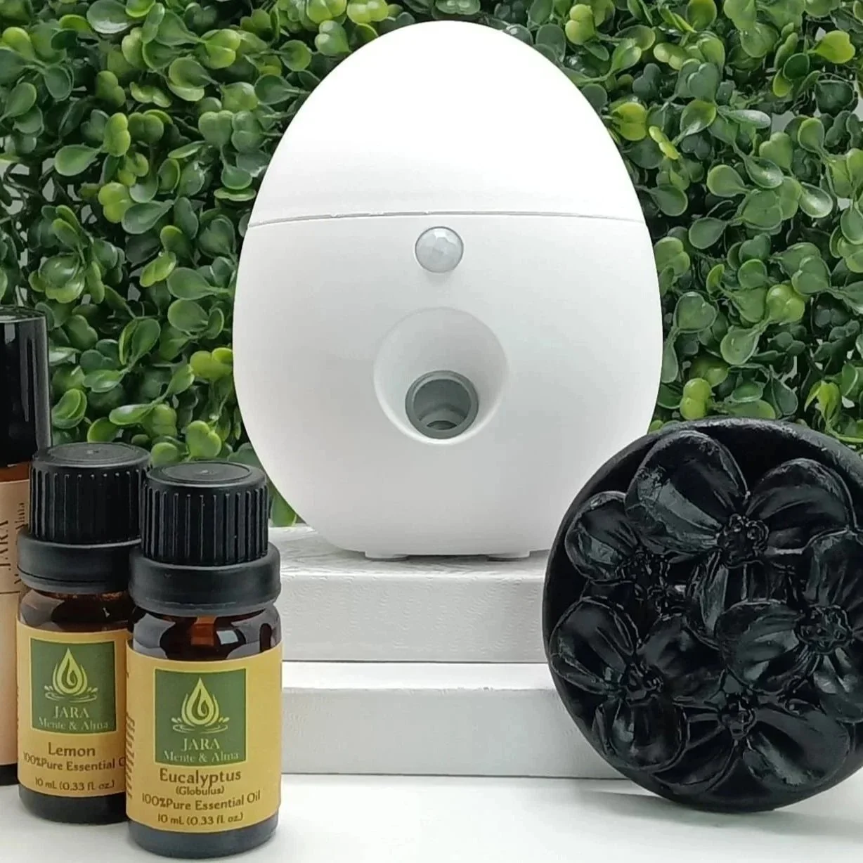Diffuser with essential oils and black soap, set against a green leafy background.
