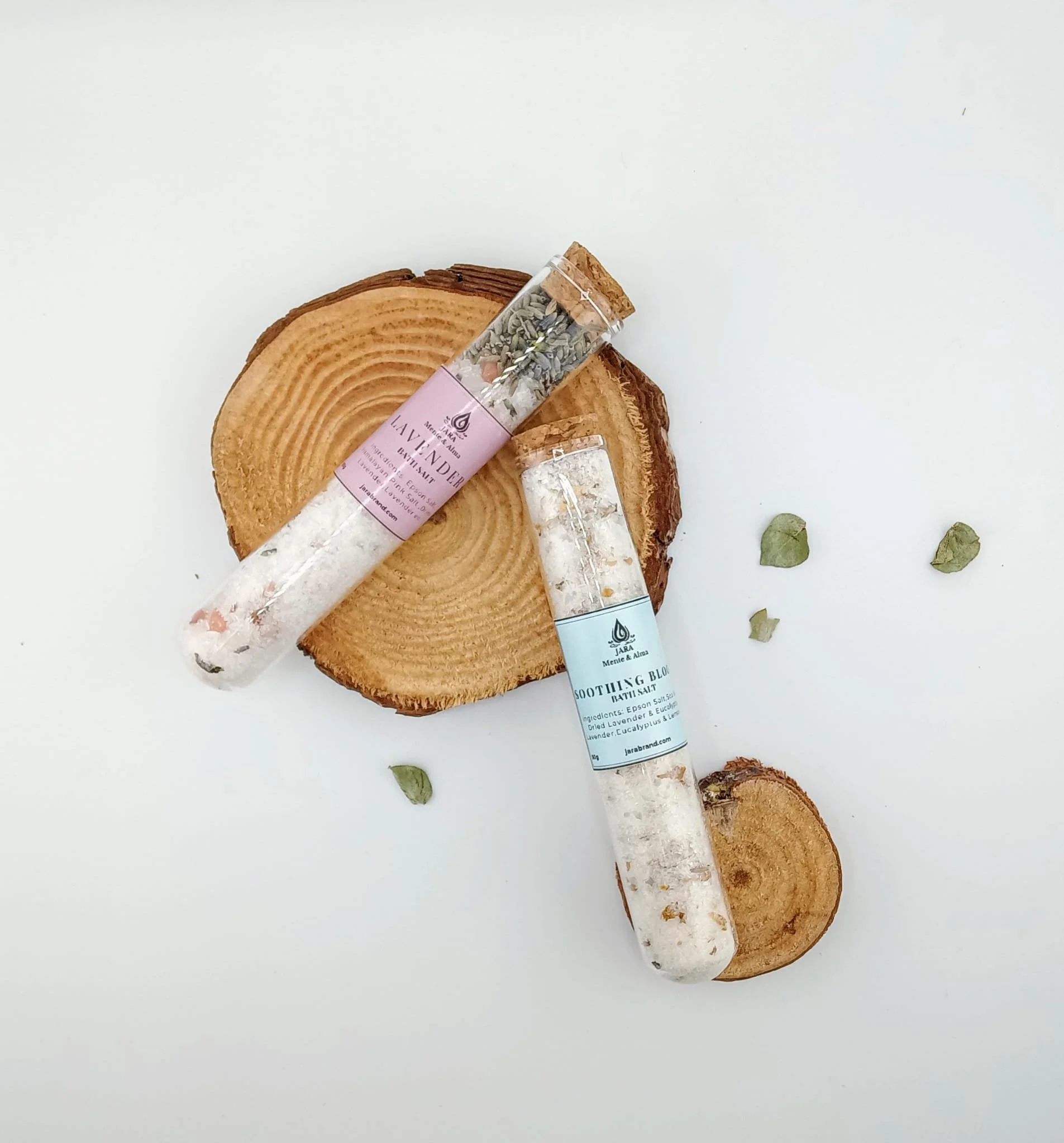 Two clear plastic tubes containing dried herbs and salts, labeled lavender and soothing balm, placed on top of two stacked wooden slices, with some small dried leaves scattered around.