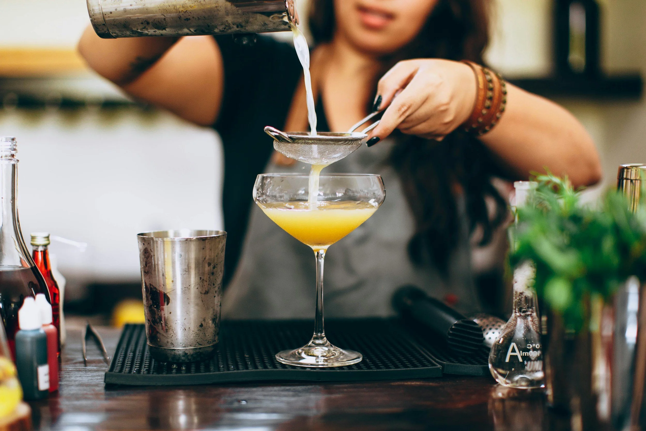 A woman pouring yellow cocktail liquid through a strainer into a cocktail glass on a bar counter, with bartending tools and bottles around.