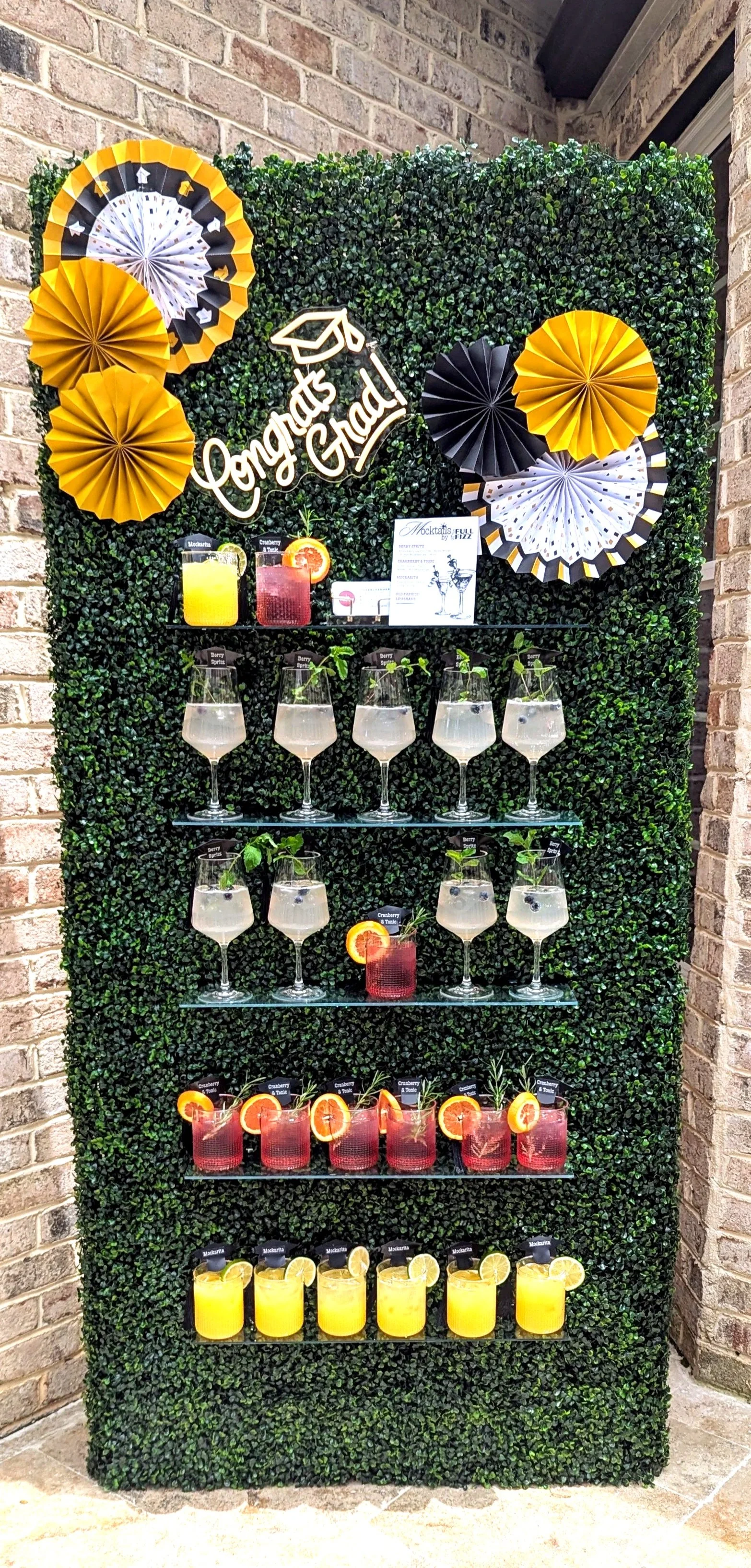 A display of cocktail glasses with garnishes on a beverage wall covered in boxwood greenery.