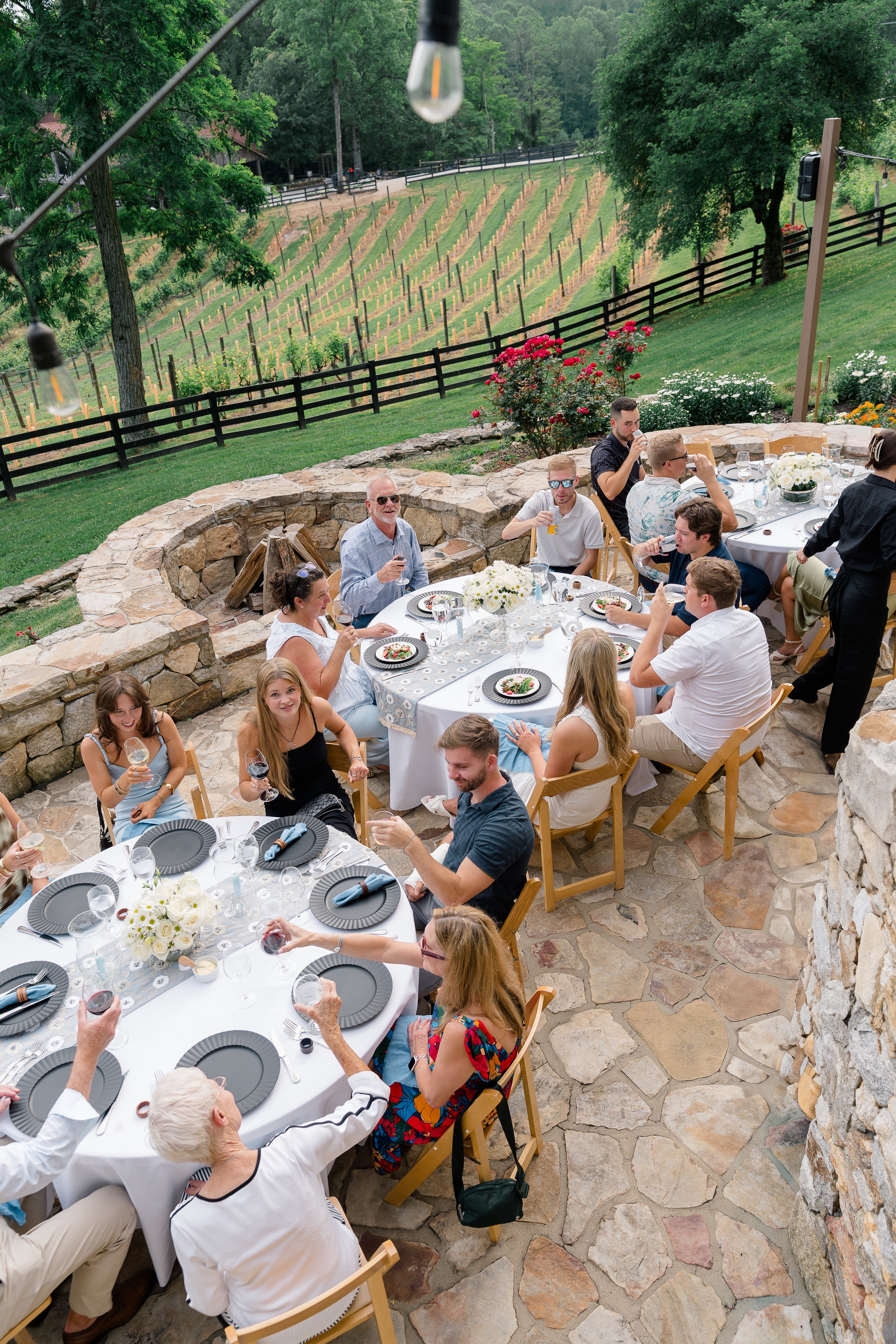 Guests enjoying a curated multi-course wine dinner at circular tables on a stone patio overlooking a vineyard.