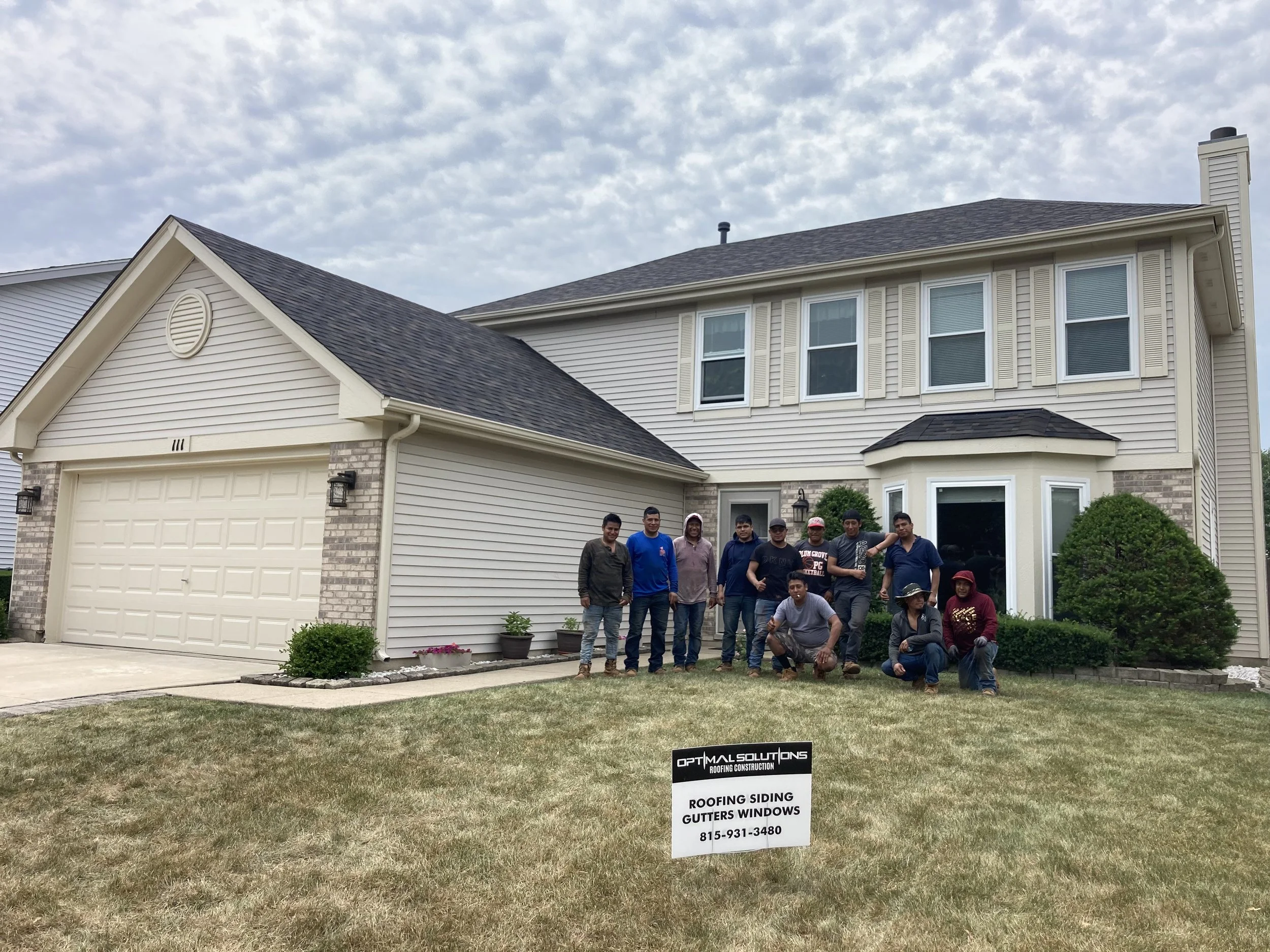A group of roofers standing in front of a residential house, with a sign in the yard advertising roofing siding gutters windows and Optimal Solutions Roofing Construction.