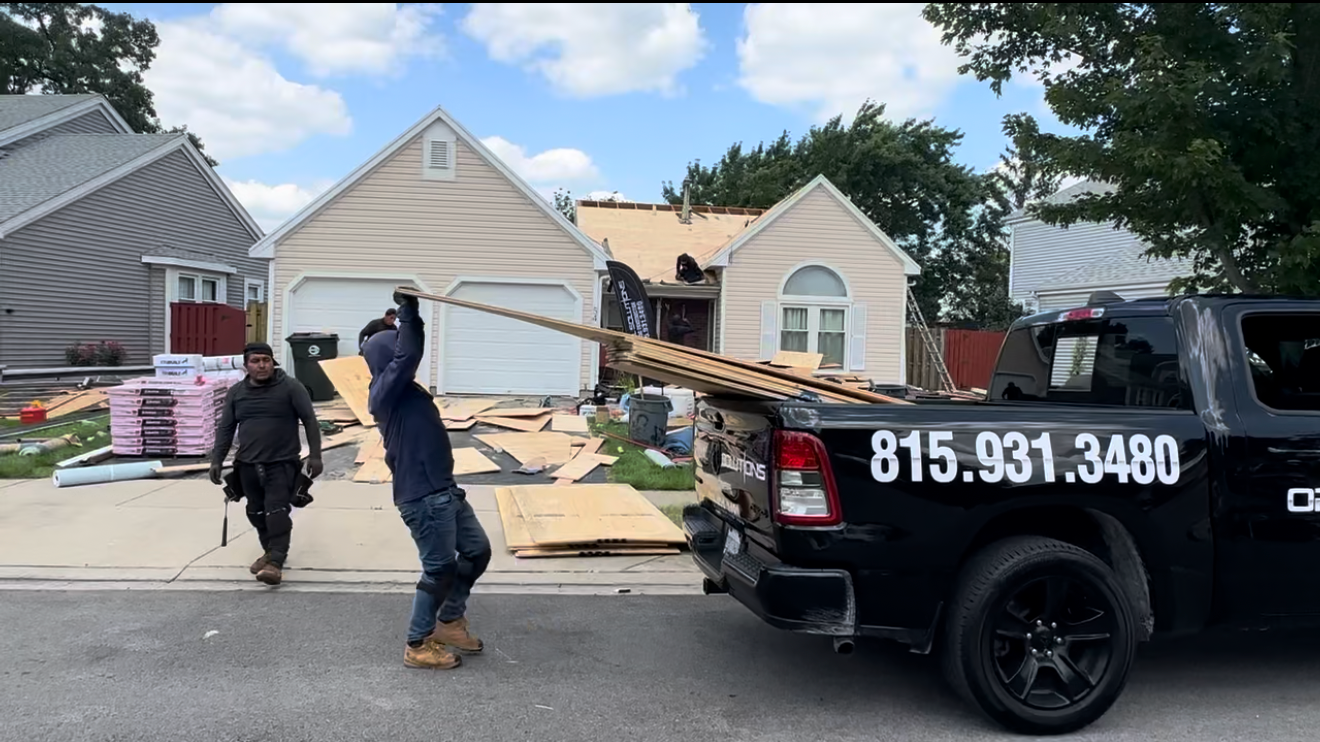 Roofing workers unloading large sheets of plywood from an Optimal Solutions truck in front of a house undergoing a new roof installation.