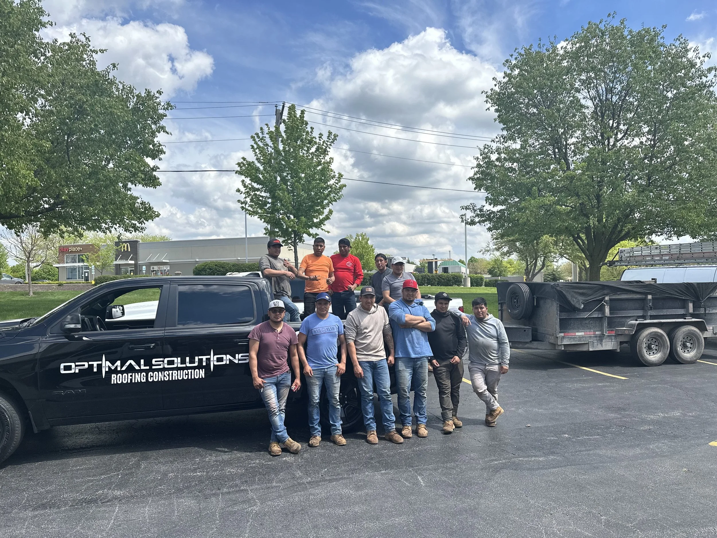 A group of twelve roofers standing next to a black pickup truck in a parking lot. The truck has a sign on the door that says "Optimum Solutions Roofing Construction."
