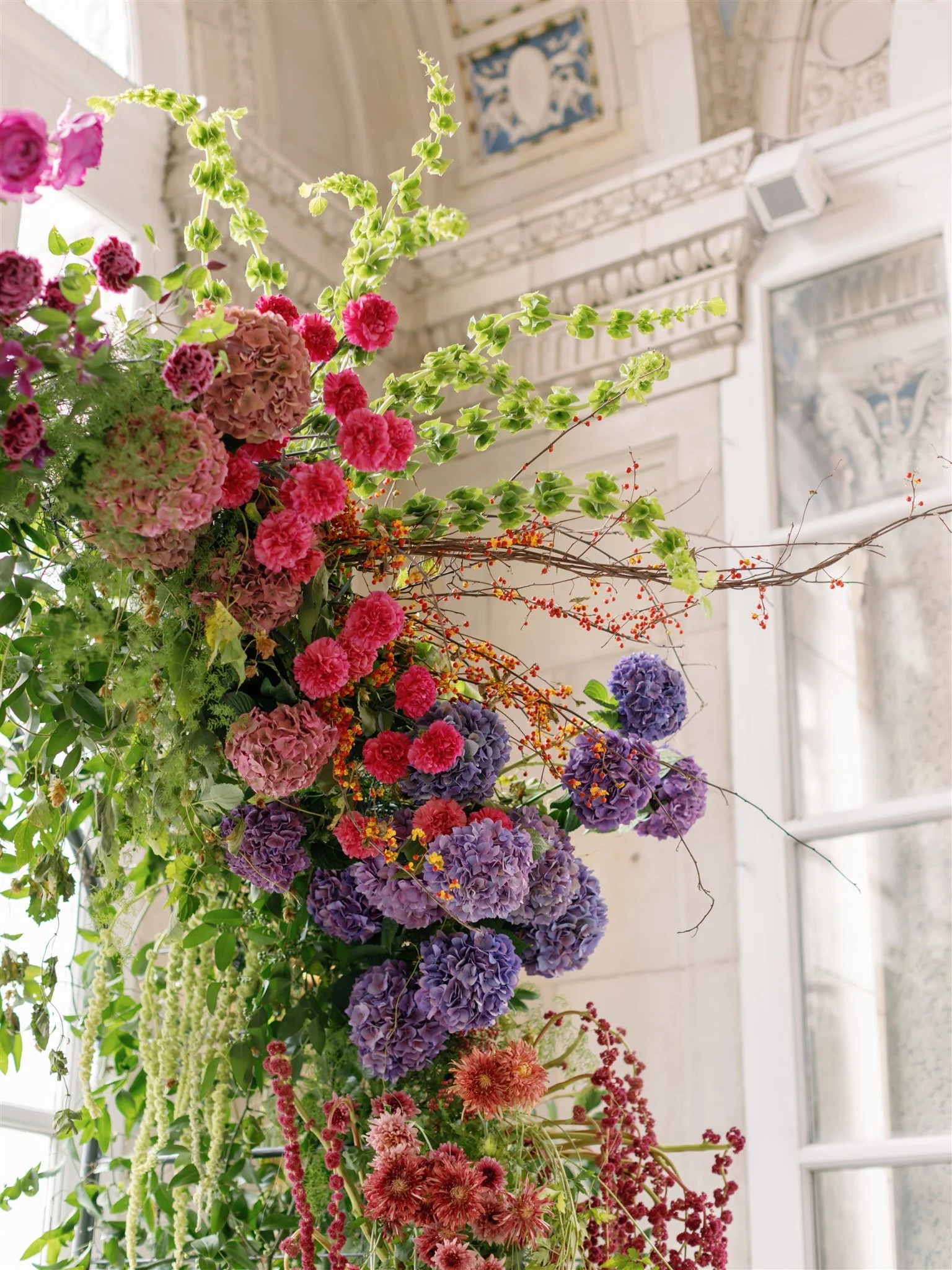 A large floral arrangement with pink, purple, and red flowers, including hydrangeas, in a bright room with intricate architectural details and large windows.