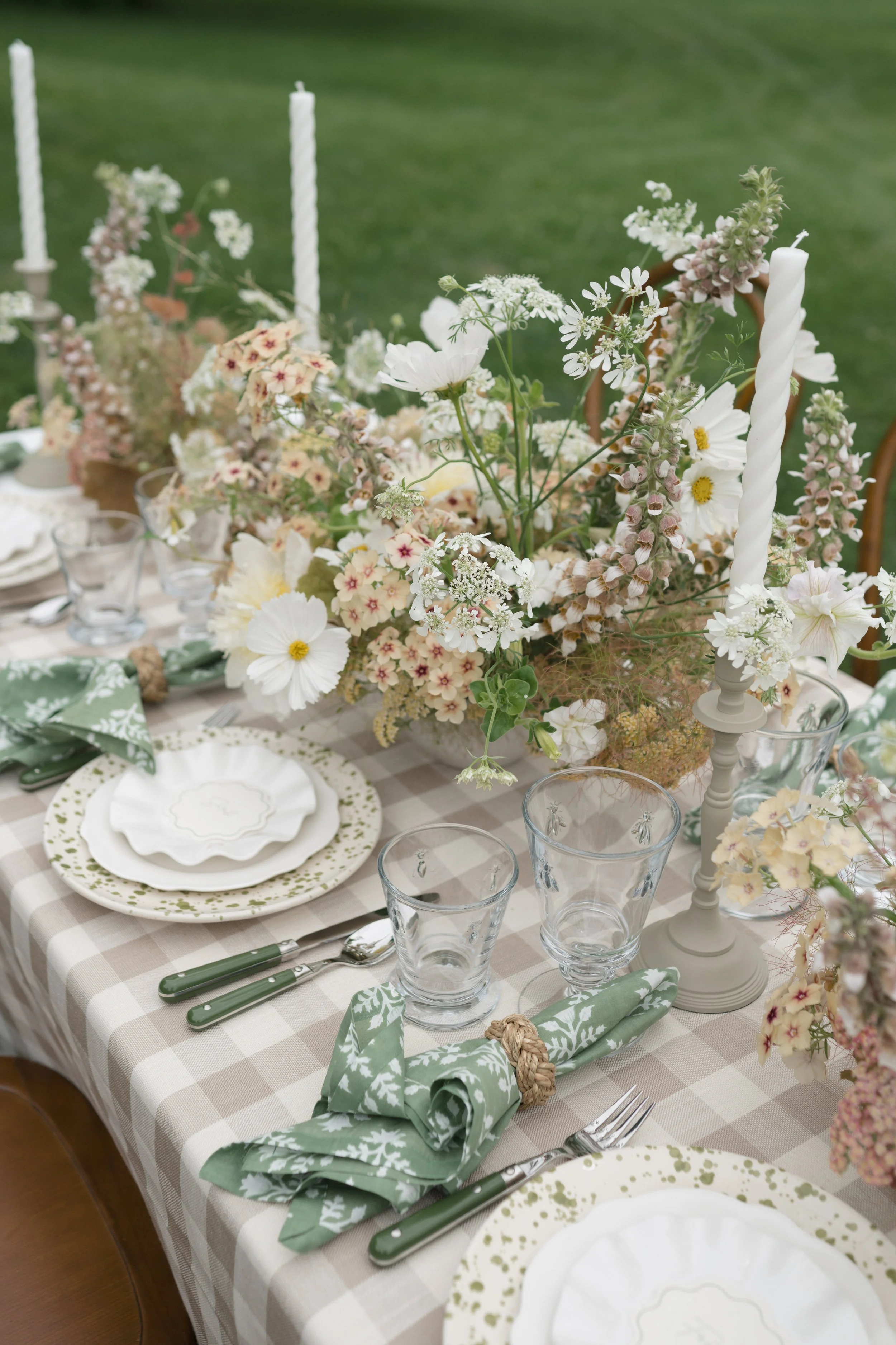 A table set for a formal outdoor event with floral arrangements, green and white patterned napkins, glassware, plates with green accents, and white candlesticks on a checkered tablecloth.