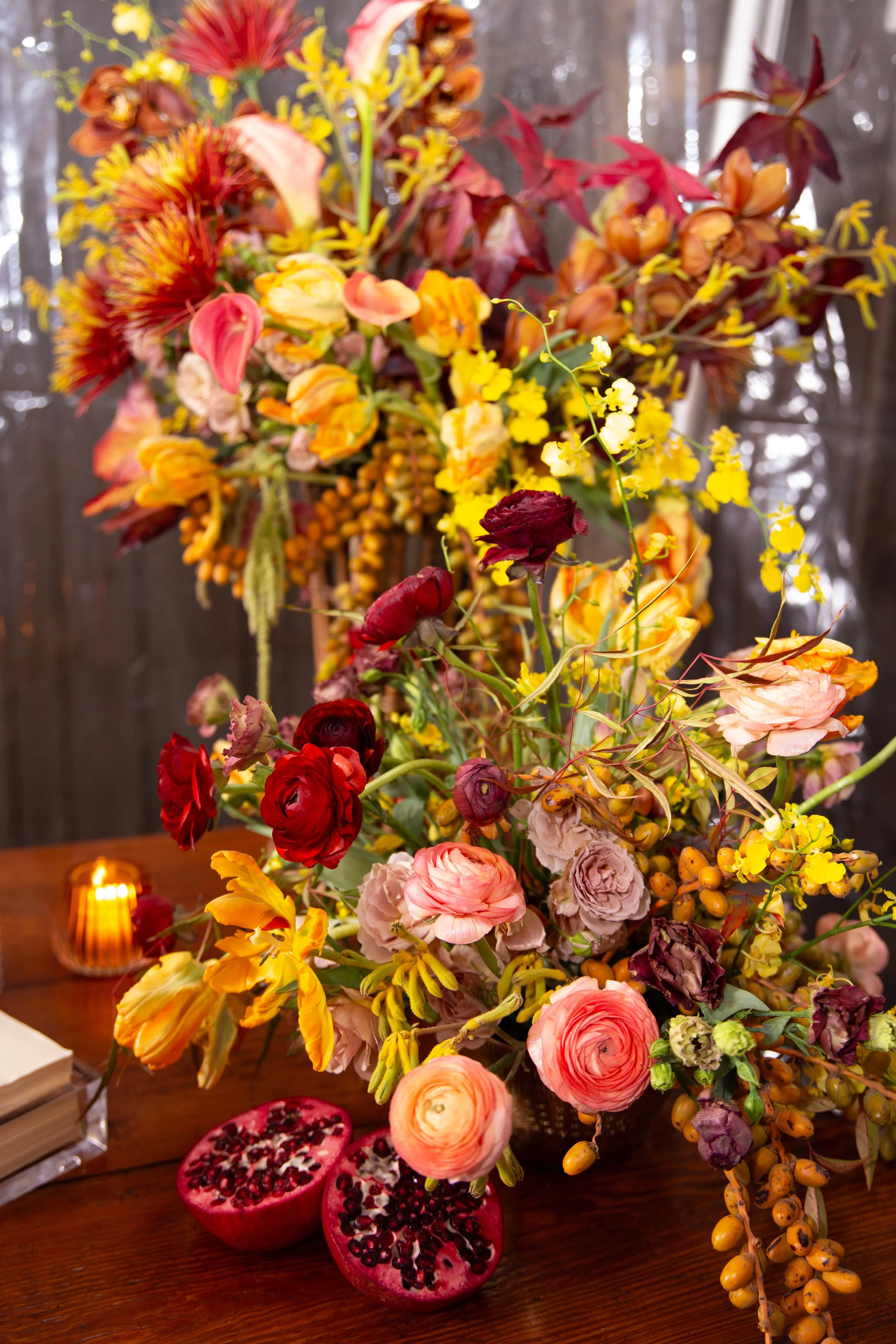 A large brightly colored floral arrangement on a wooden table includes pink, red, yellow, and purple flowers with a bunch of berries. Two halves of a pomegranate are placed in front of the flowers, and a small lit candle is in the background.