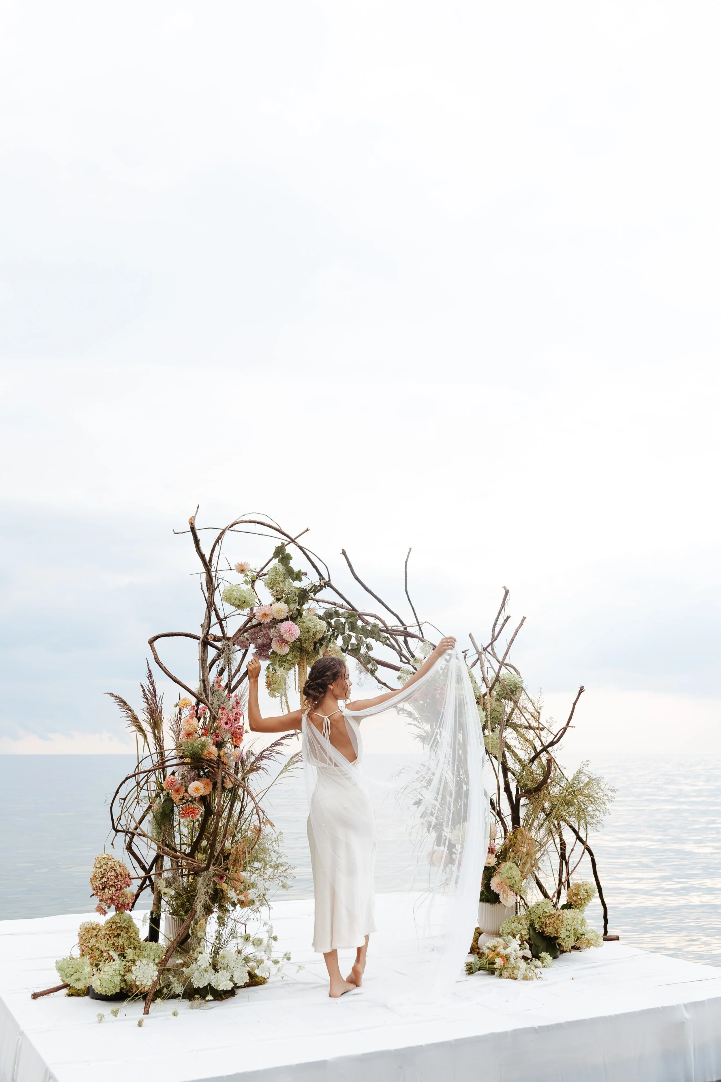 WNY Barcelona, NY A woman in a white dress standing on a decorated platform by the water, holding a sheer fabric, with an arch made of twigs, flowers, and greenery behind her.