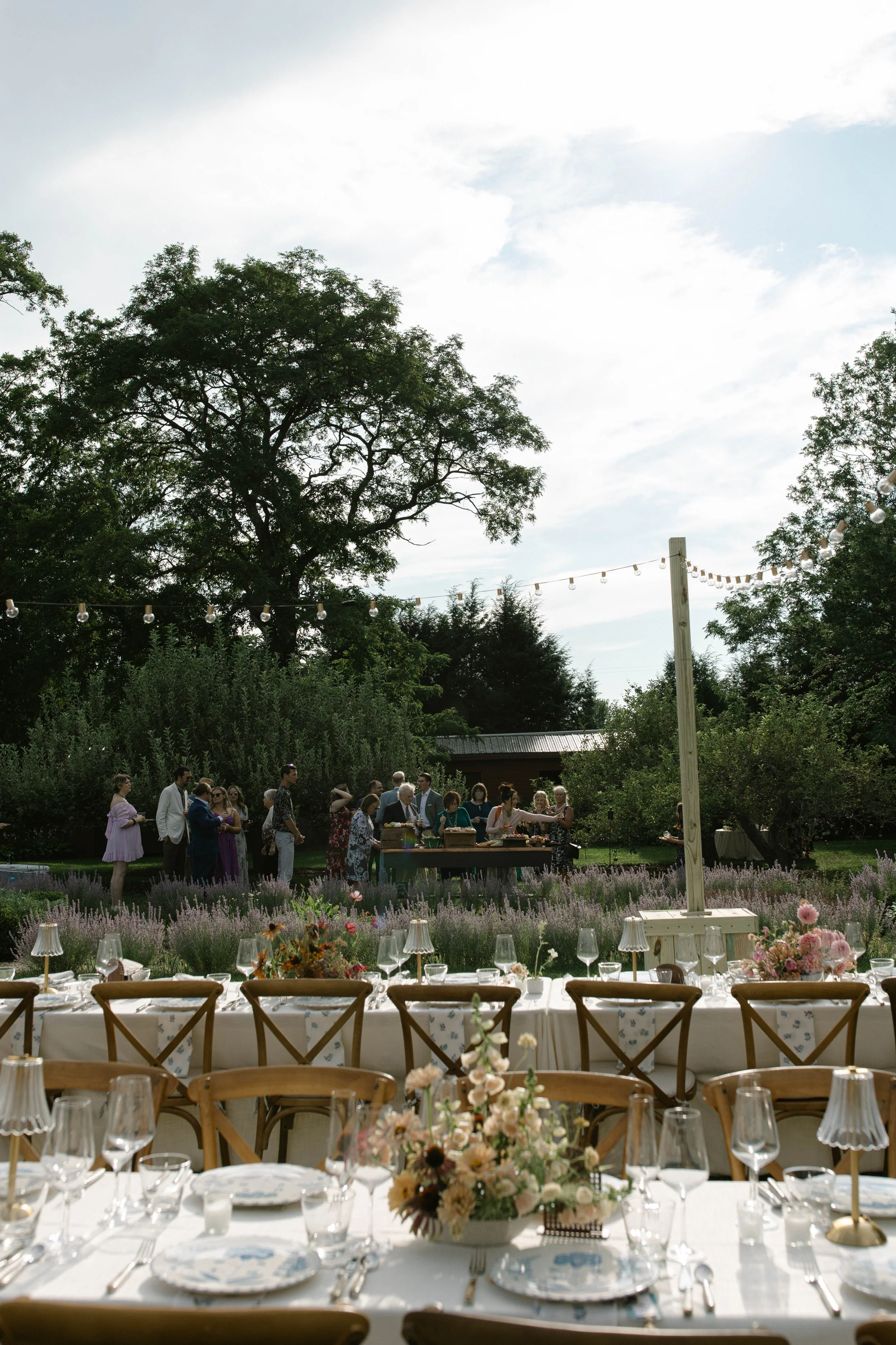 Johnson Estate Winery Westfield, NY An outdoor event setup with a long dining table decorated with floral centerpieces and a celebratory gathering of people in formal attire in the background under string lights in a garden setting.