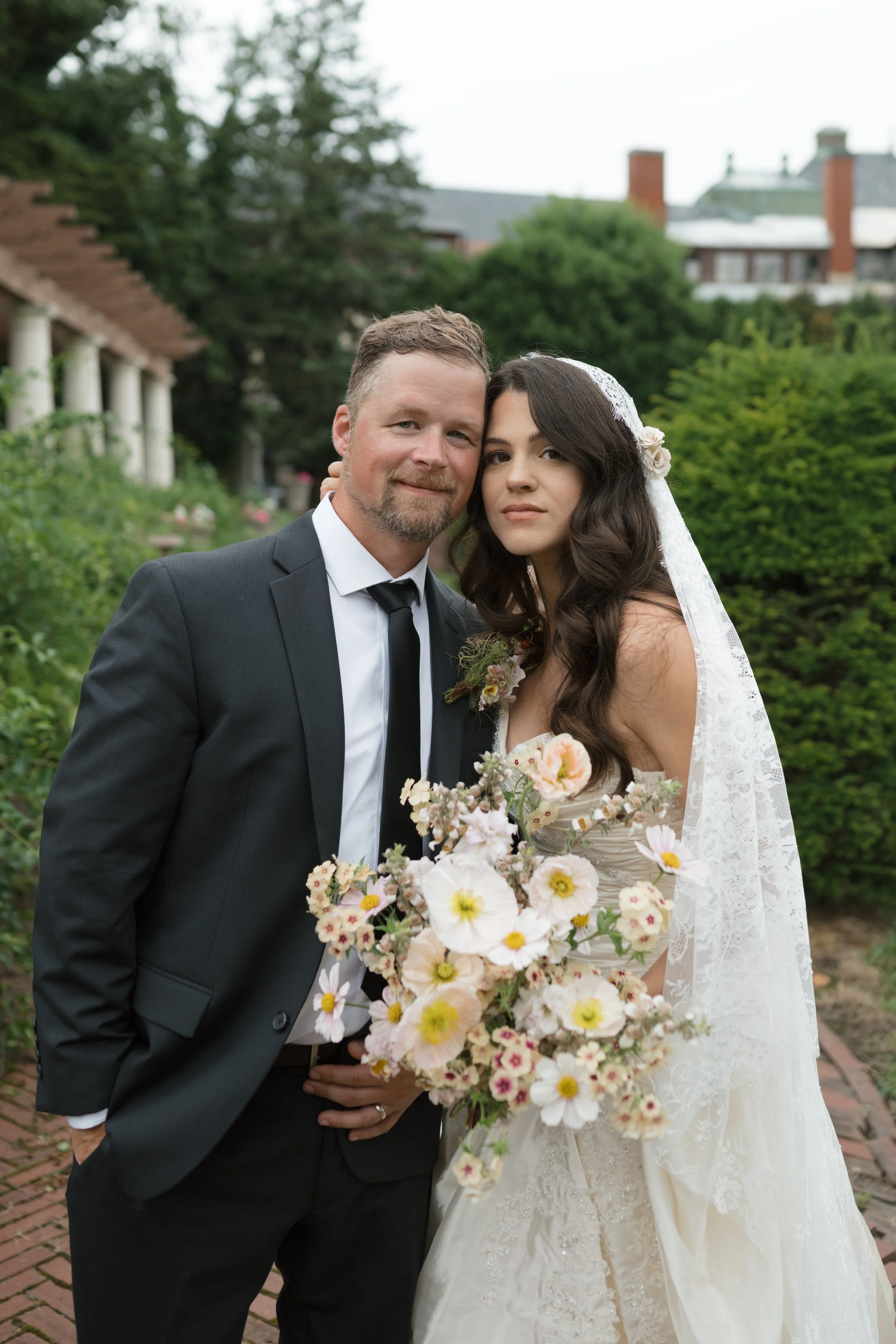 A bride and groom pose together outdoors on a wedding day, holding a bouquet of pink and white flowers. The bride wears a white lace wedding gown and veil, while the groom is dressed in a black suit with a white shirt and black tie. They stand in a garden with greenery and a brick pathway.