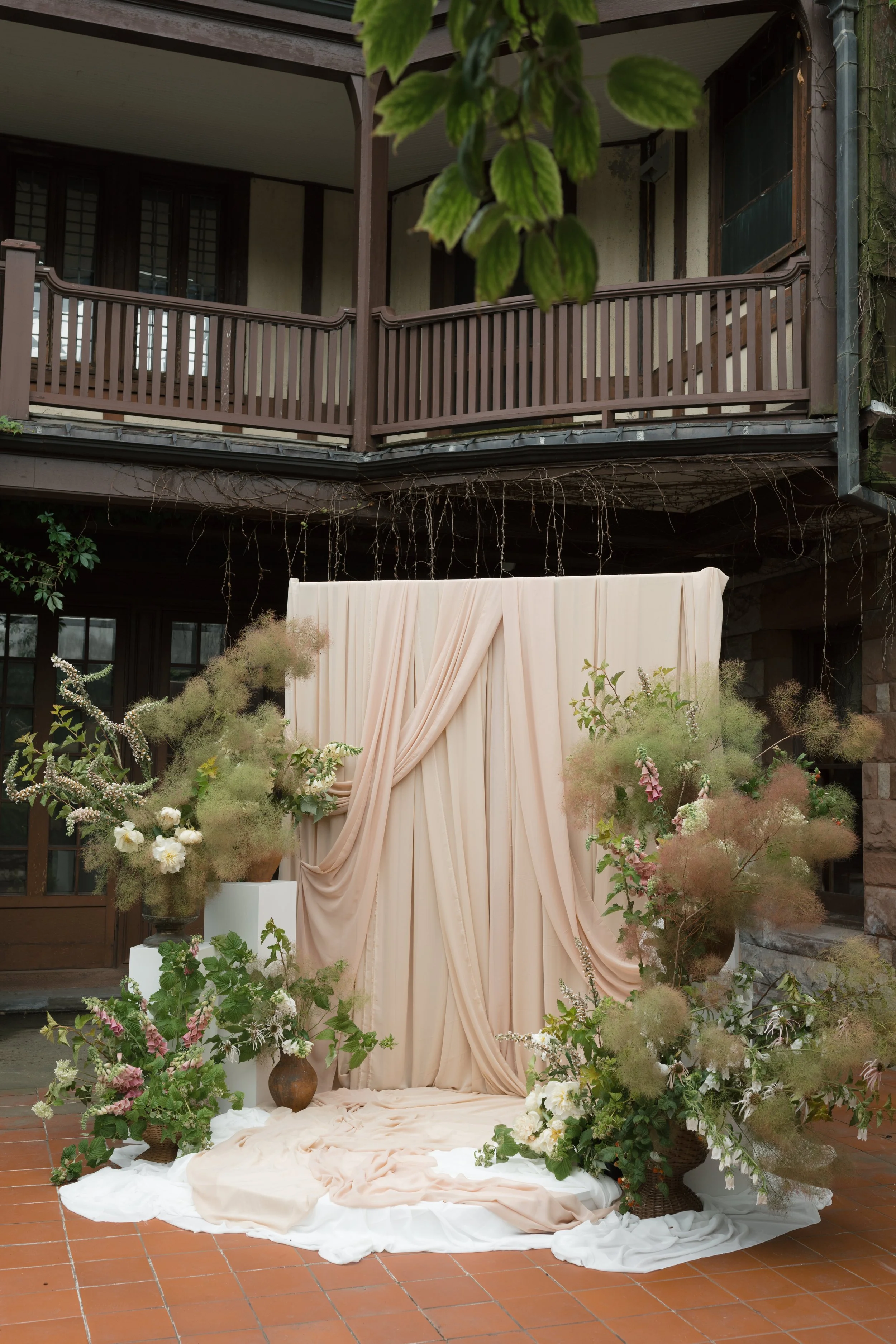 Sonnenberg Gardens, Canandaigua, NY A decorative setup with beige draped fabric and lush floral arrangements, likely for an outdoor event or wedding, on a tiled patio with a wooden balcony above.