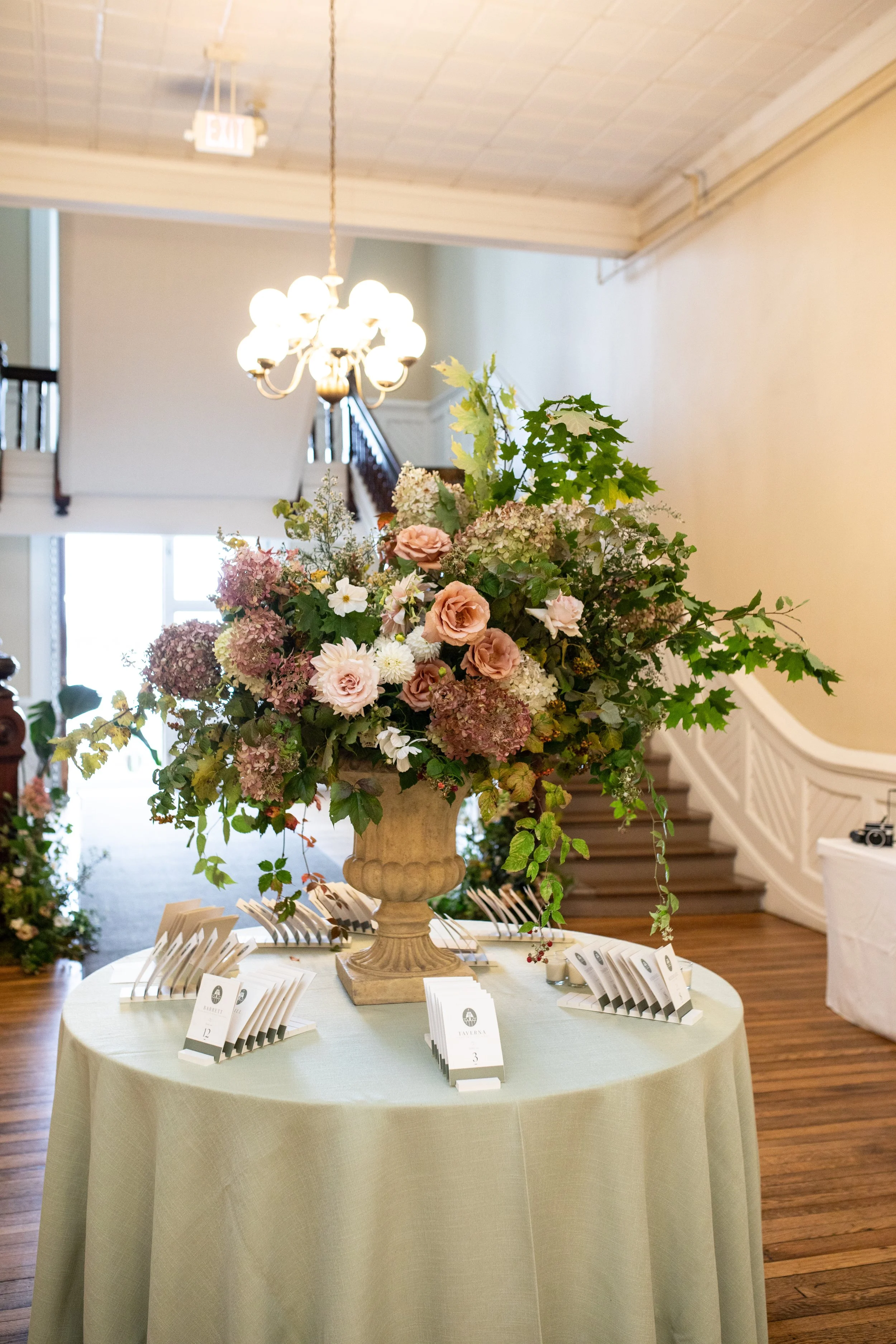 Chautauqua Institution wedding A large floral arrangement with pink and white flowers in a stone vase on a round table covered with a light green tablecloth at an indoor event.