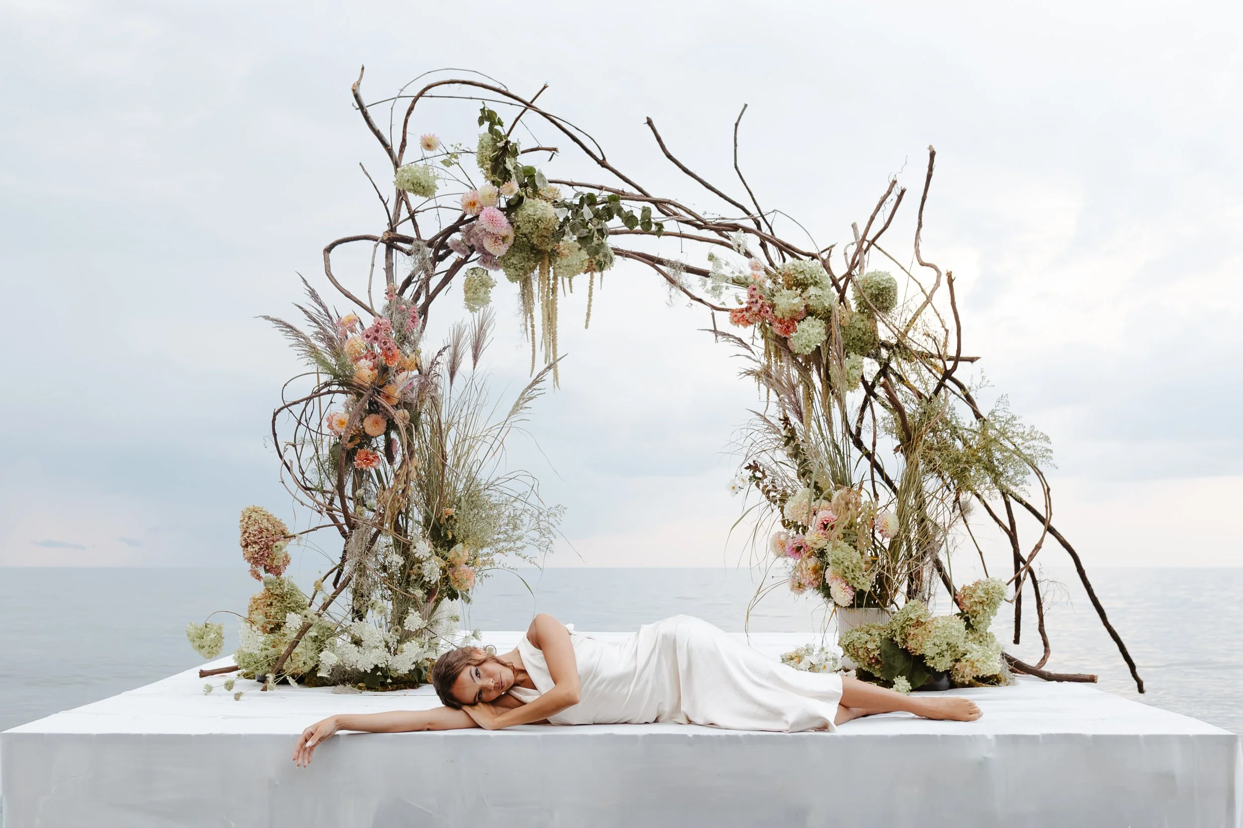 WNY - Barcelona, NY creative content collaboration. A woman lying down on a platform in front of a decorative floral arch by the ocean.