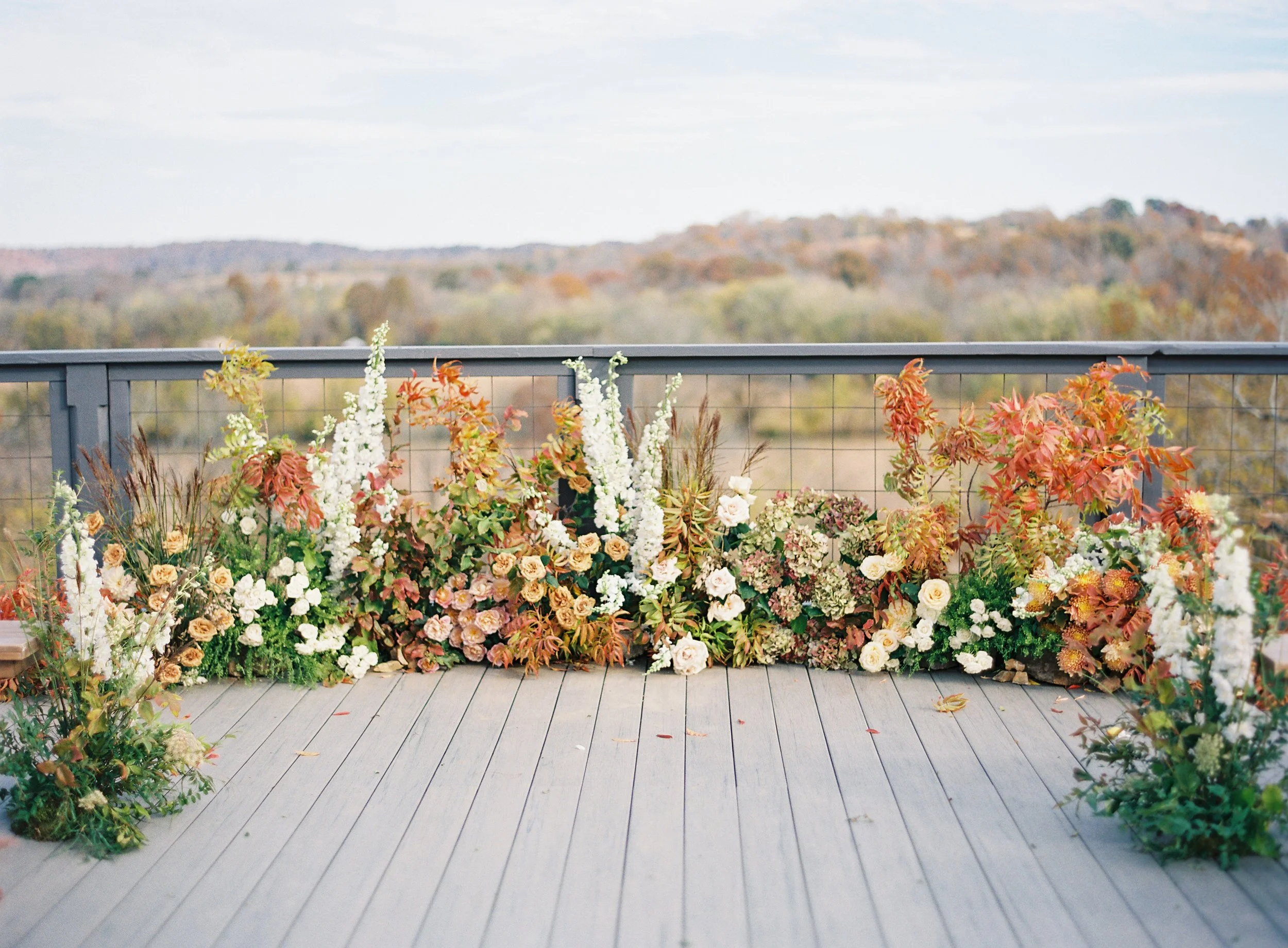 Outdoor balcony with a wooden floor and a railing, decorated with various colorful flowers and plants, with a scenic view of a distant landscape and trees in autumn colors.