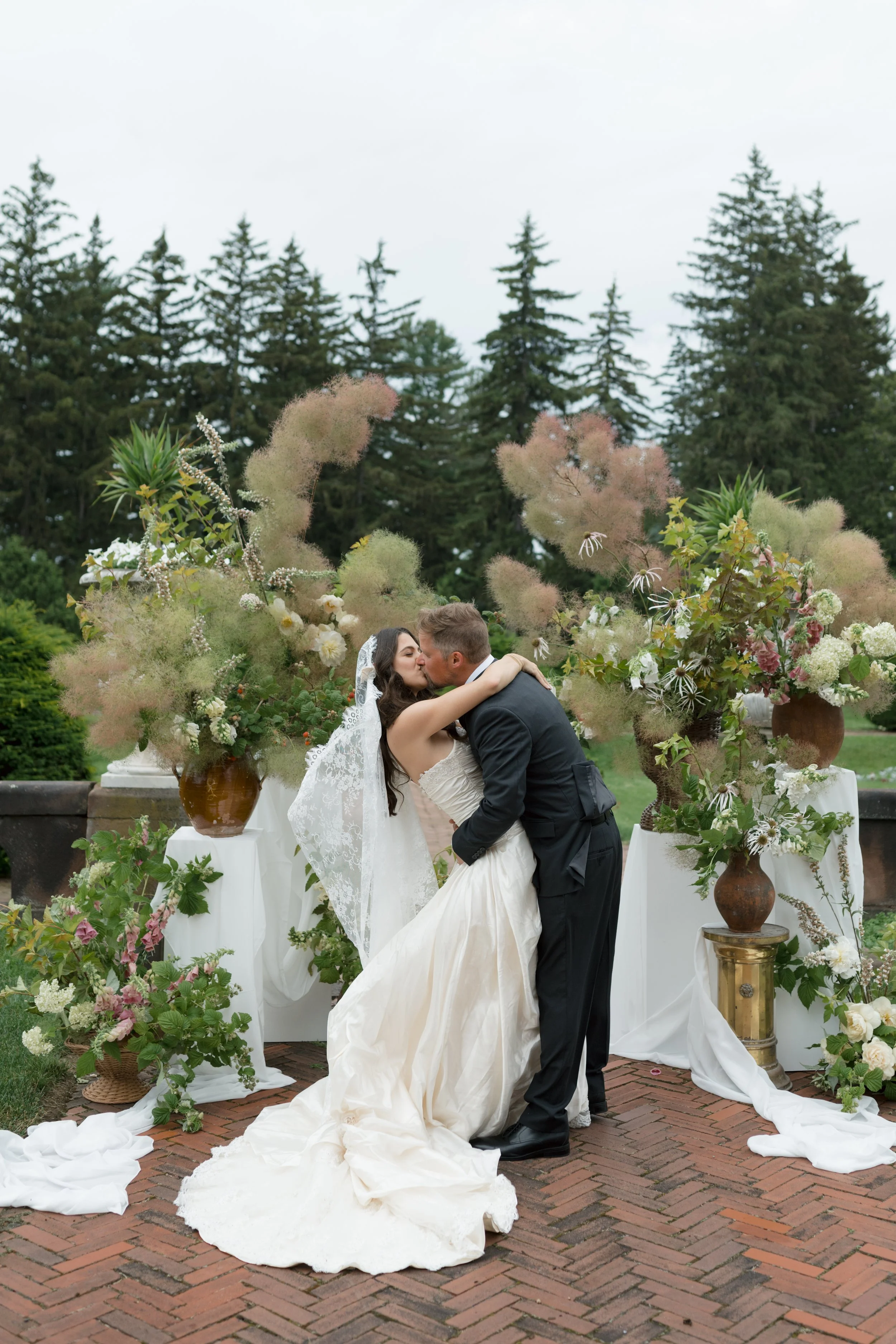 Sonnenberg Gardens, Canandaigua, NY. Bride and groom sharing a kiss surrounded by large floral arrangements on stands, outdoors on a brick patio during a cloudy day.