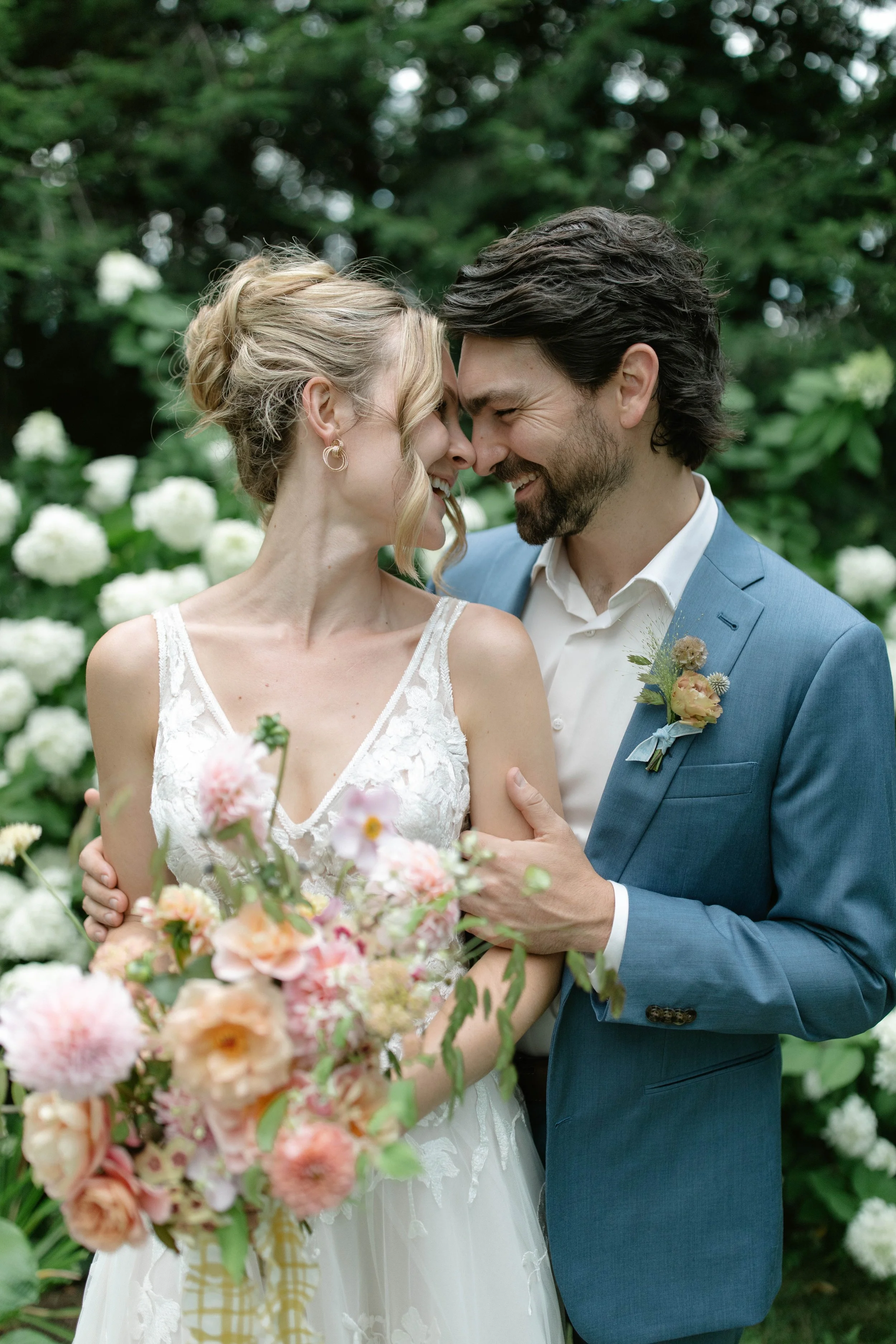 A bride and groom in wedding attire sharing a close, joyful moment outdoors with lush greenery and white flowering bushes in the background.