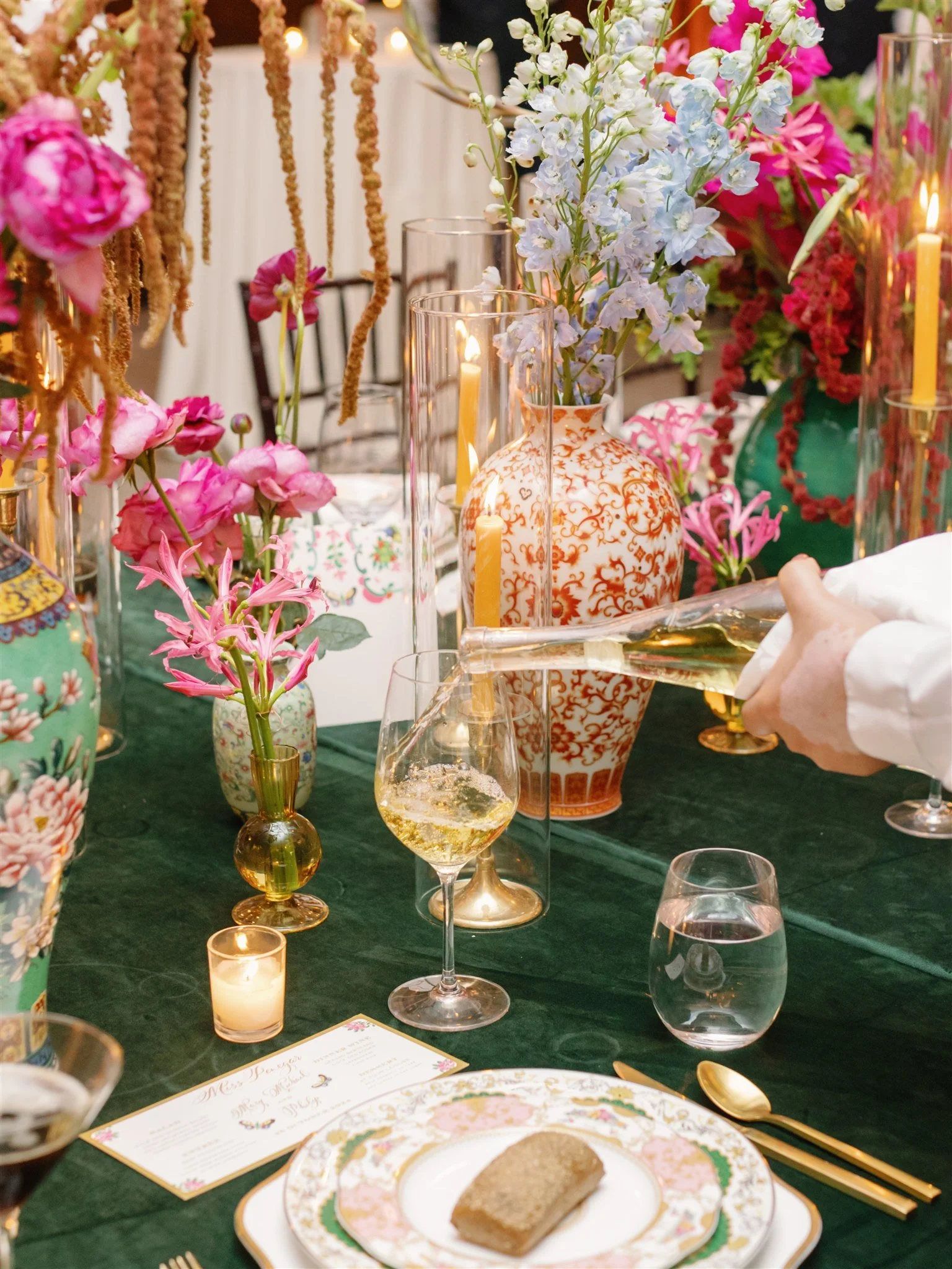 A table decorated with colorful flowers, vases, lit candles, and a hand pouring a drink. Plate with a slice of cake, utensils, and a menu are also visible.