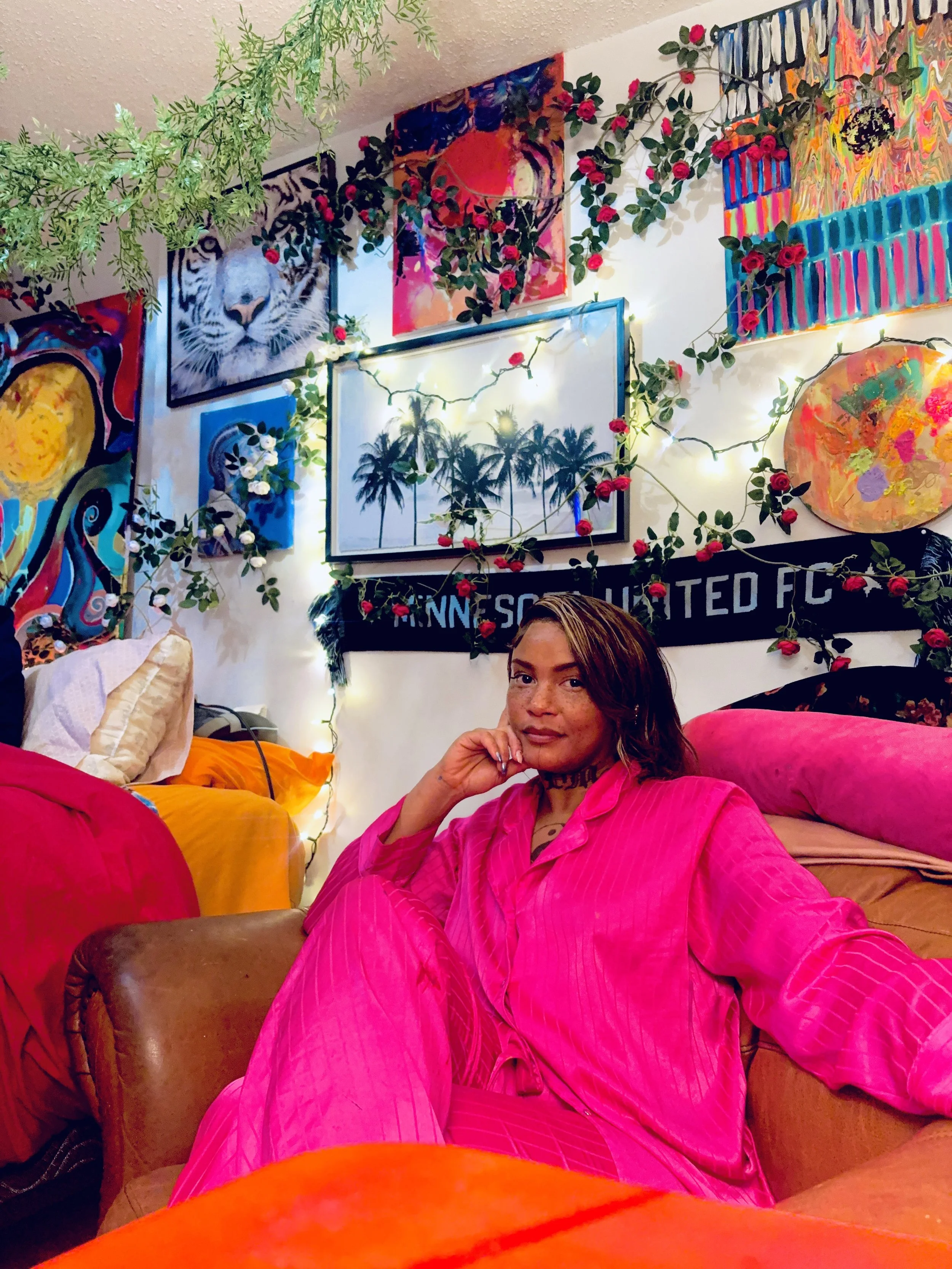 A woman with long brown hair and freckles sitting on a pink couch in a colorful room decorated with artwork, string lights, and greenery.