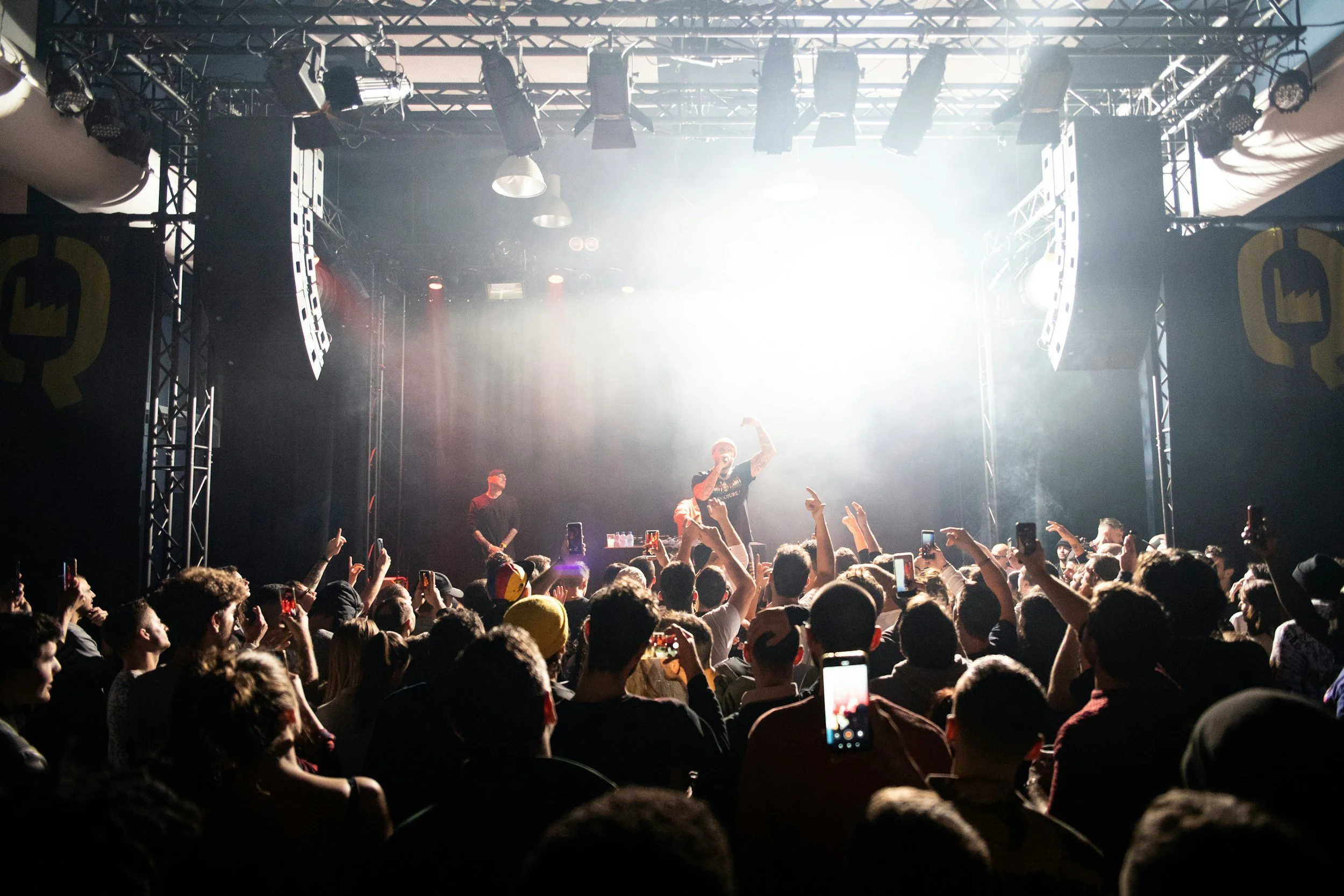 A concert stage with bright lights and a crowd of people, some holding up their phones, watching a performer on stage.
