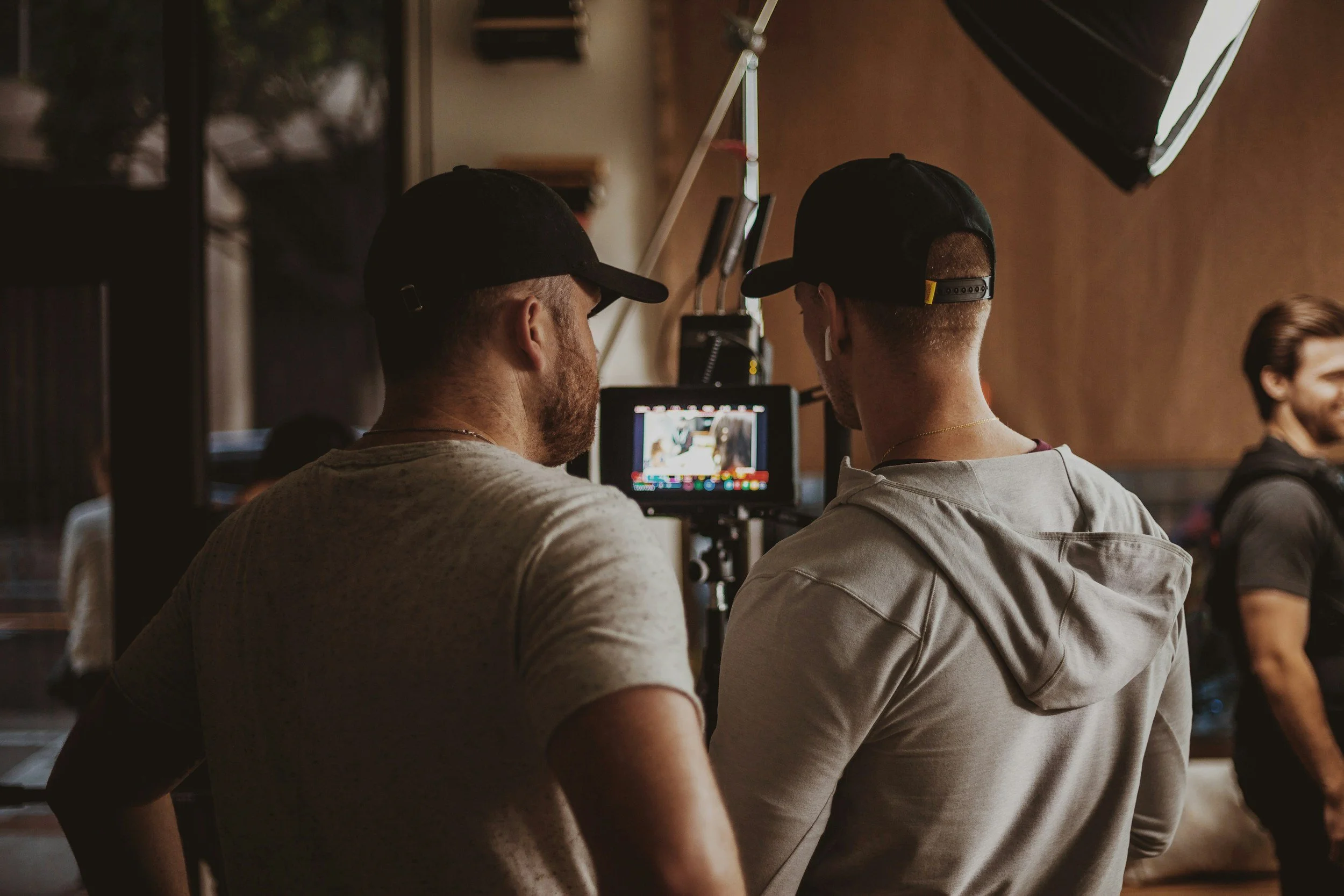 Two men wearing black caps and gray shirts are looking at a camera monitor on a tripod in a room with warm lighting, with a woman smiling in the background.