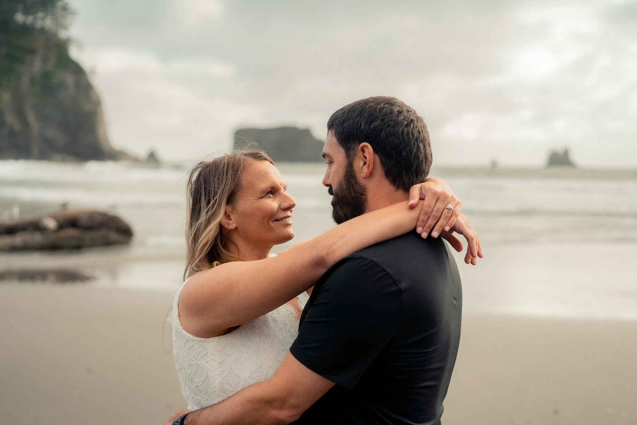 A couple embracing on the beach with ocean waves and cliffs in the background, looking into each other's eyes.