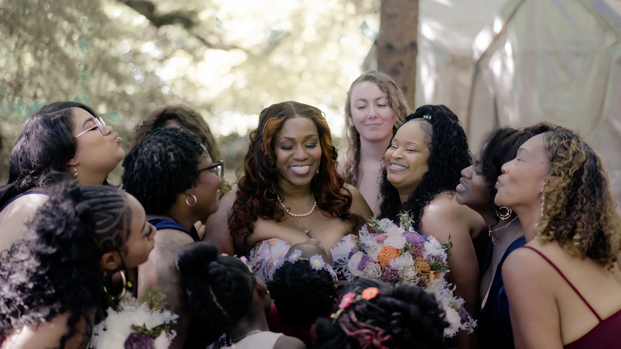 A group of women, with diverse hairstyles and skin tones, gathered outdoors in a circle, smiling and embracing, one woman holding a bouquet of flowers amidst sunlight filtering through trees.