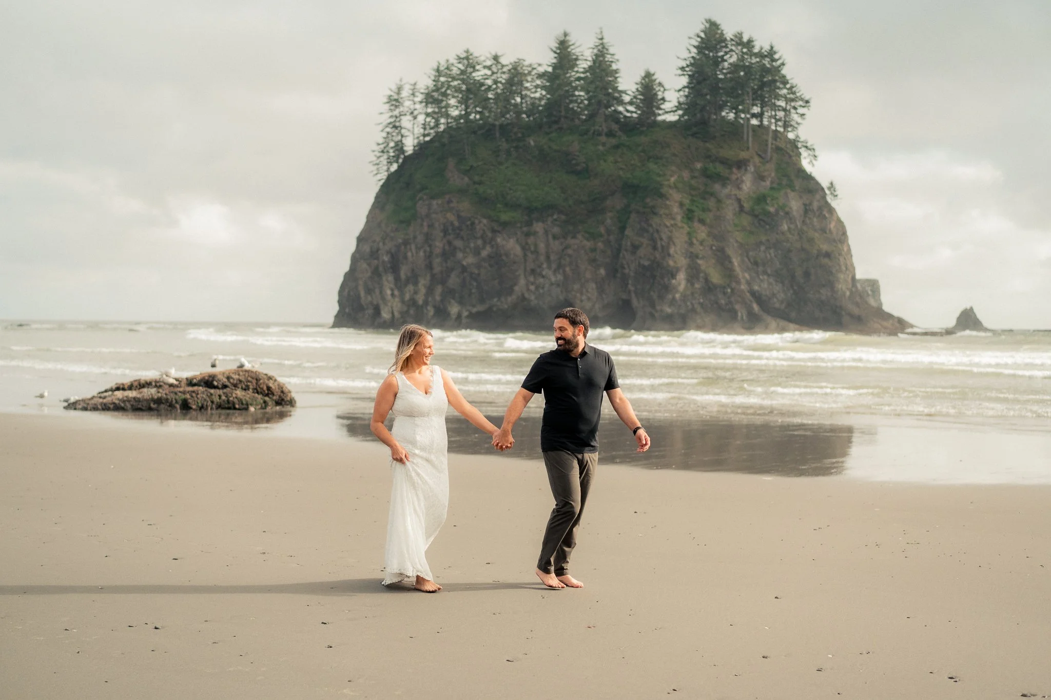 A couple holding hands walking on a beach with a large rocky island covered with trees in the background.