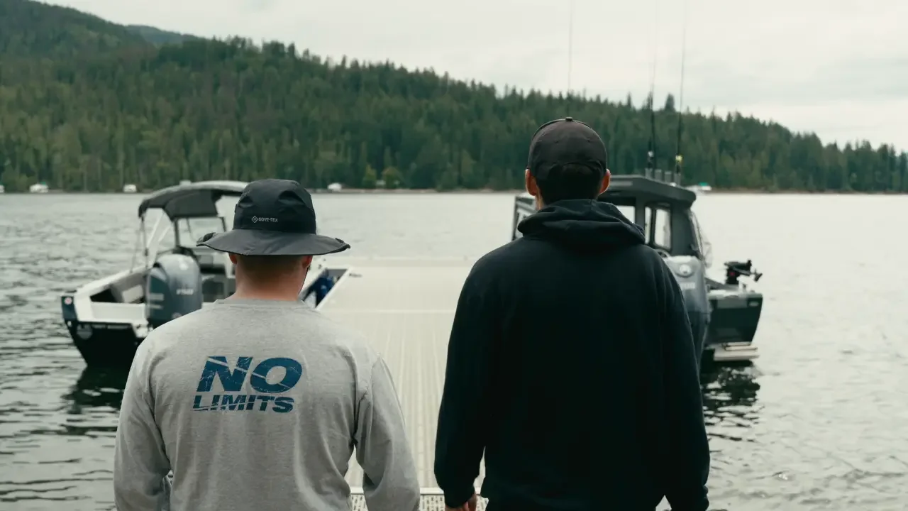 Two men stand on a dock, looking at boats on a lake with a forested shoreline and overcast sky in the background.