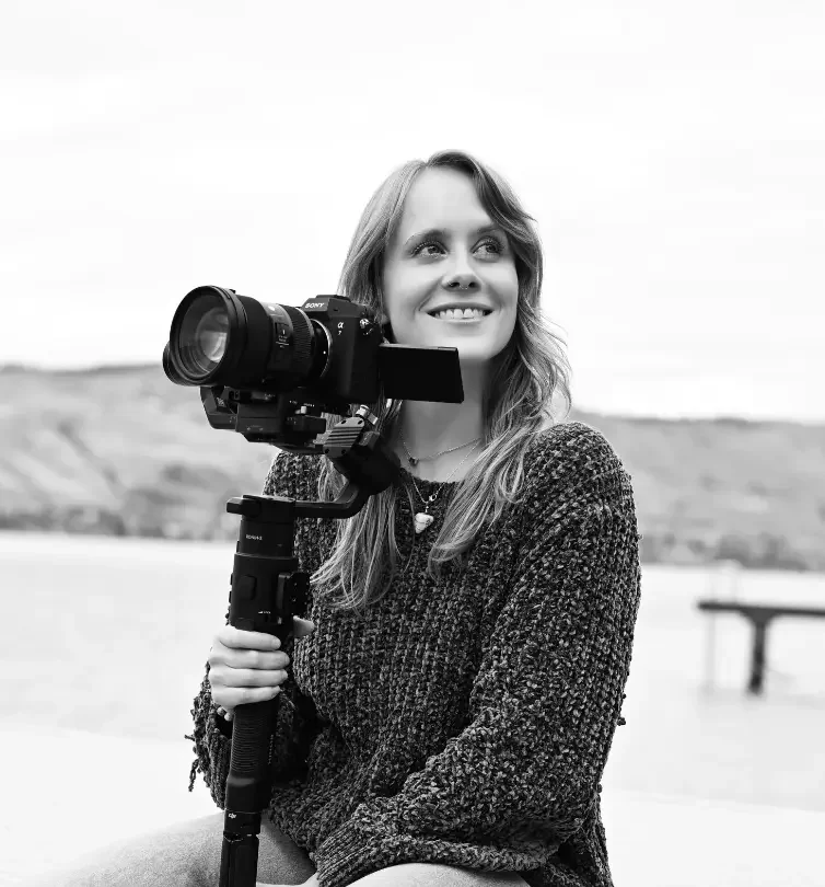 A woman holding a professional video camera on a tripod outdoors, smiling and looking upward, with a blurred landscape in the background.