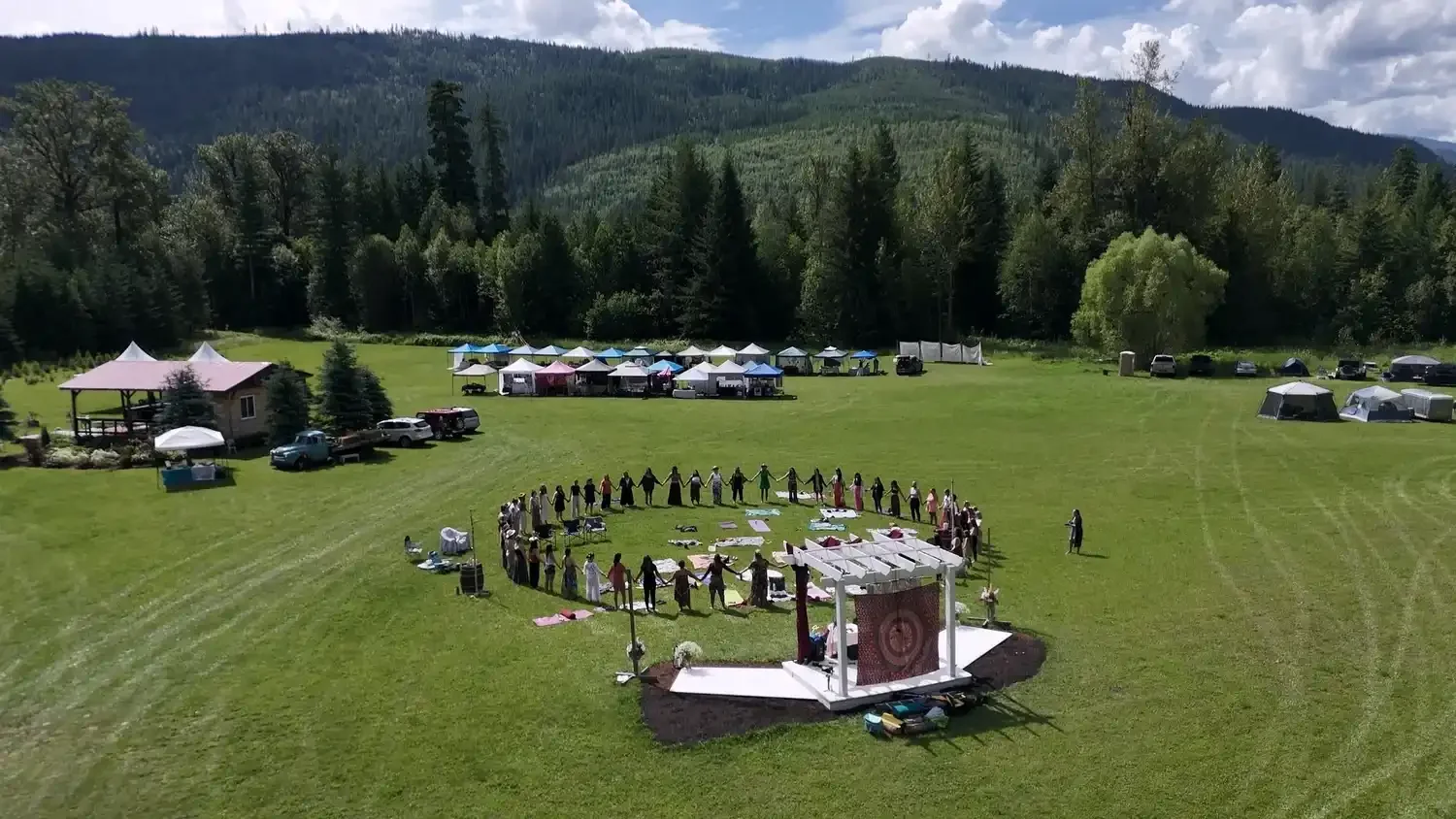 A group of people holding hands in a circle outdoors on a grassy field, with tents and vehicles in the background, mountains and trees in the distance.