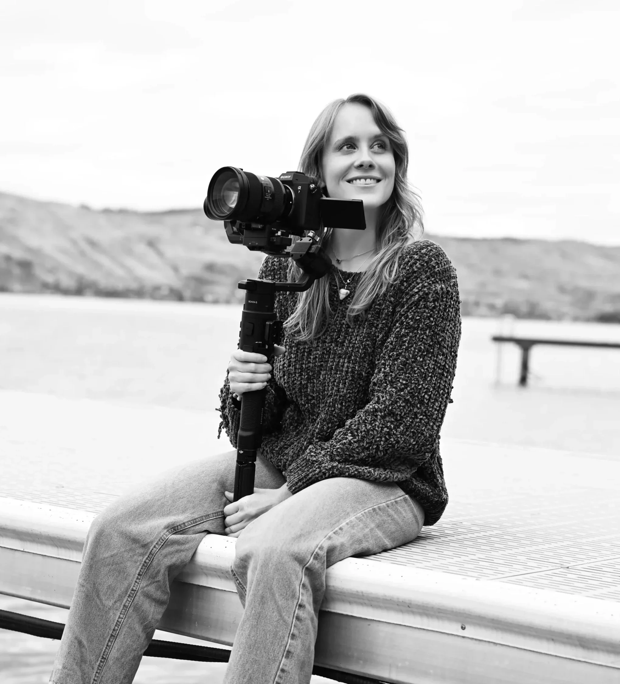 A young woman sitting on a boat dock, smiling with a camera on her shoulder, dressed in casual clothes, with a blurred outdoor background.