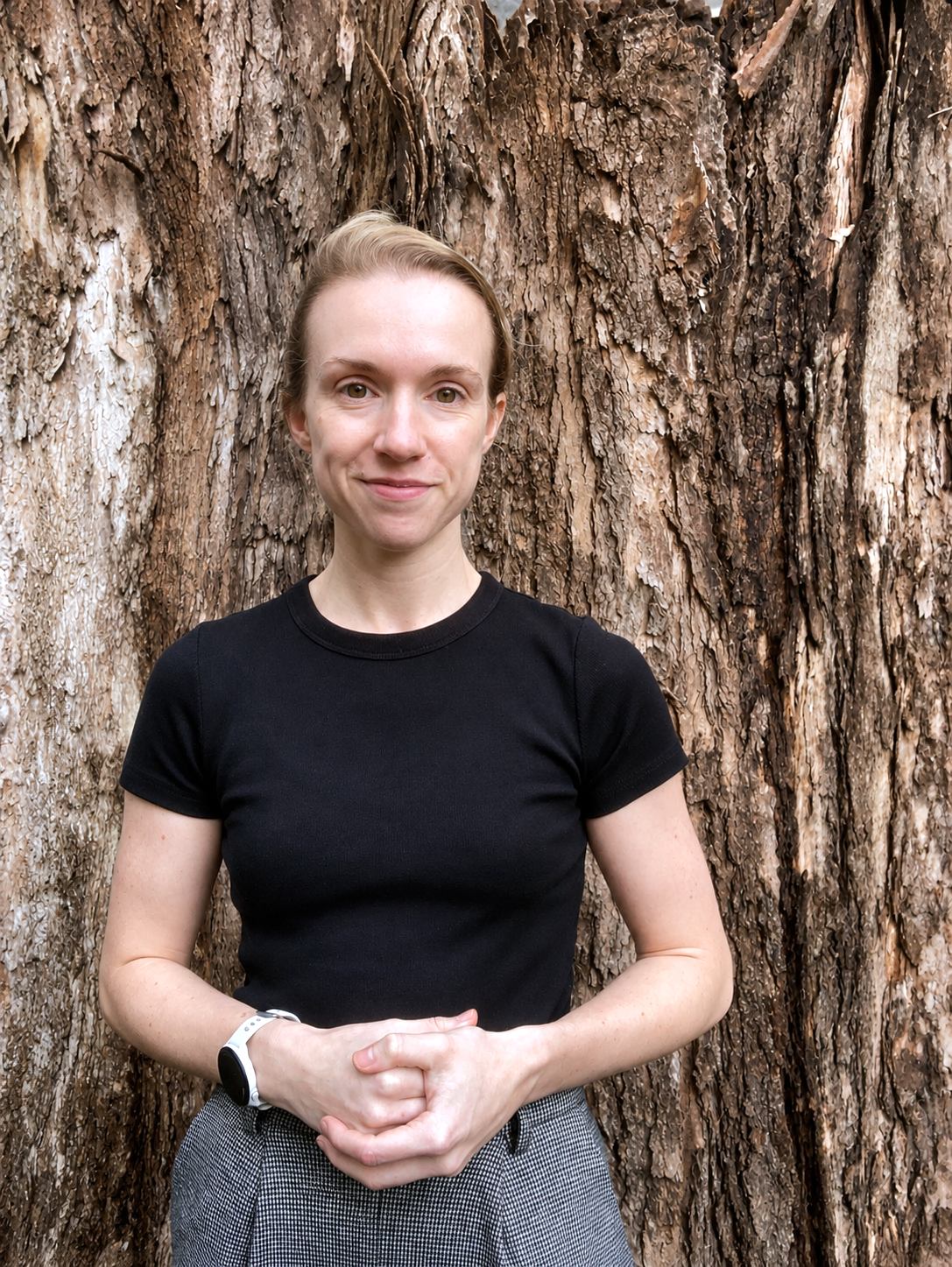 A woman stands in front of a tree form waist height, smiling with her hands clasped