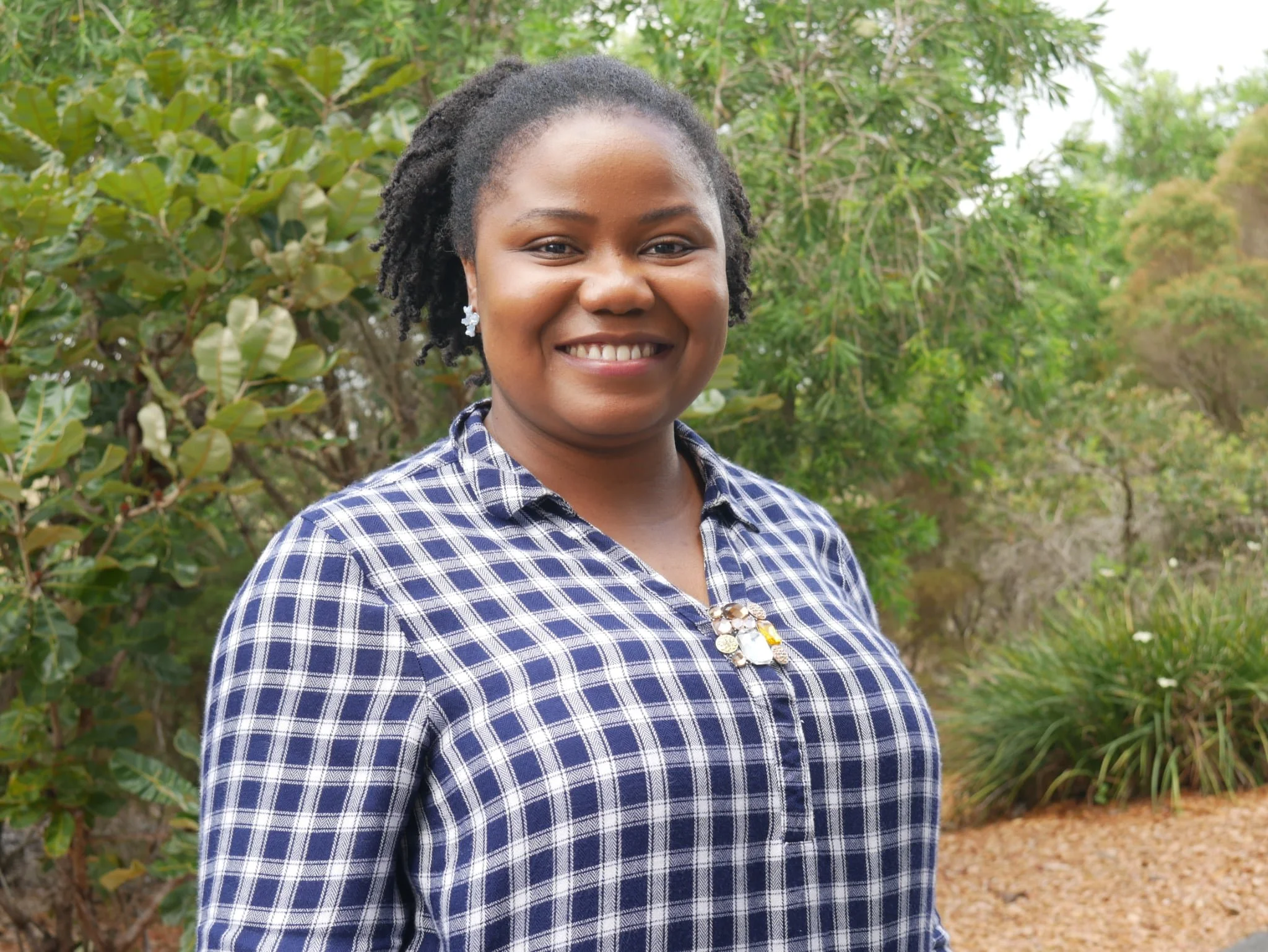 Chioma Ojiako, Youth Group Leader, Portrait of a smiling woman with dark hair styled in twists, wearing a blue and white checkered shirt and star-shaped earrings, standing outdoors in front of green foliage.