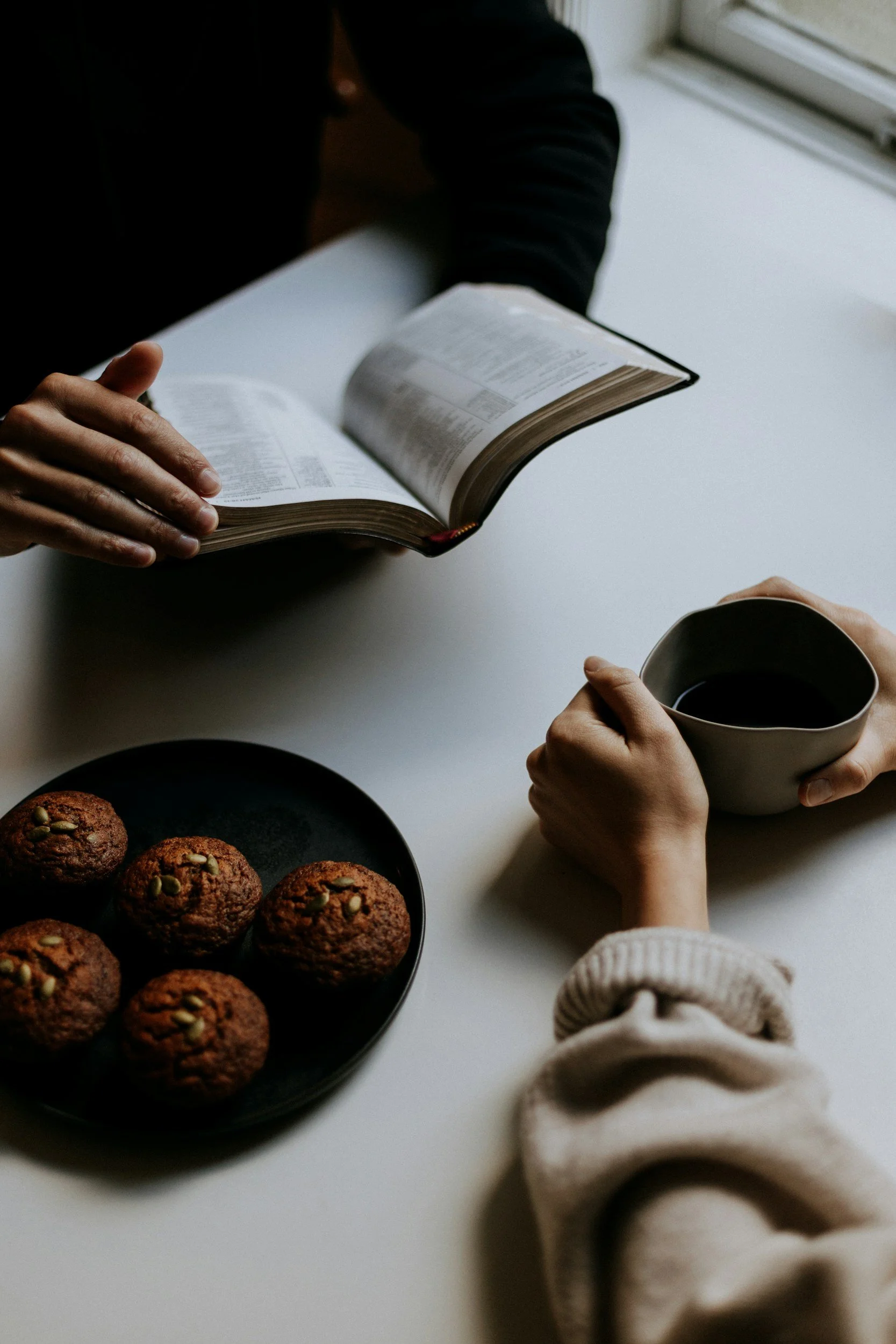 People sitting around a white table with a black plate of muffins, a coffee mug, and an open book. One person is holding a mug, and another is reaching for a muffin.