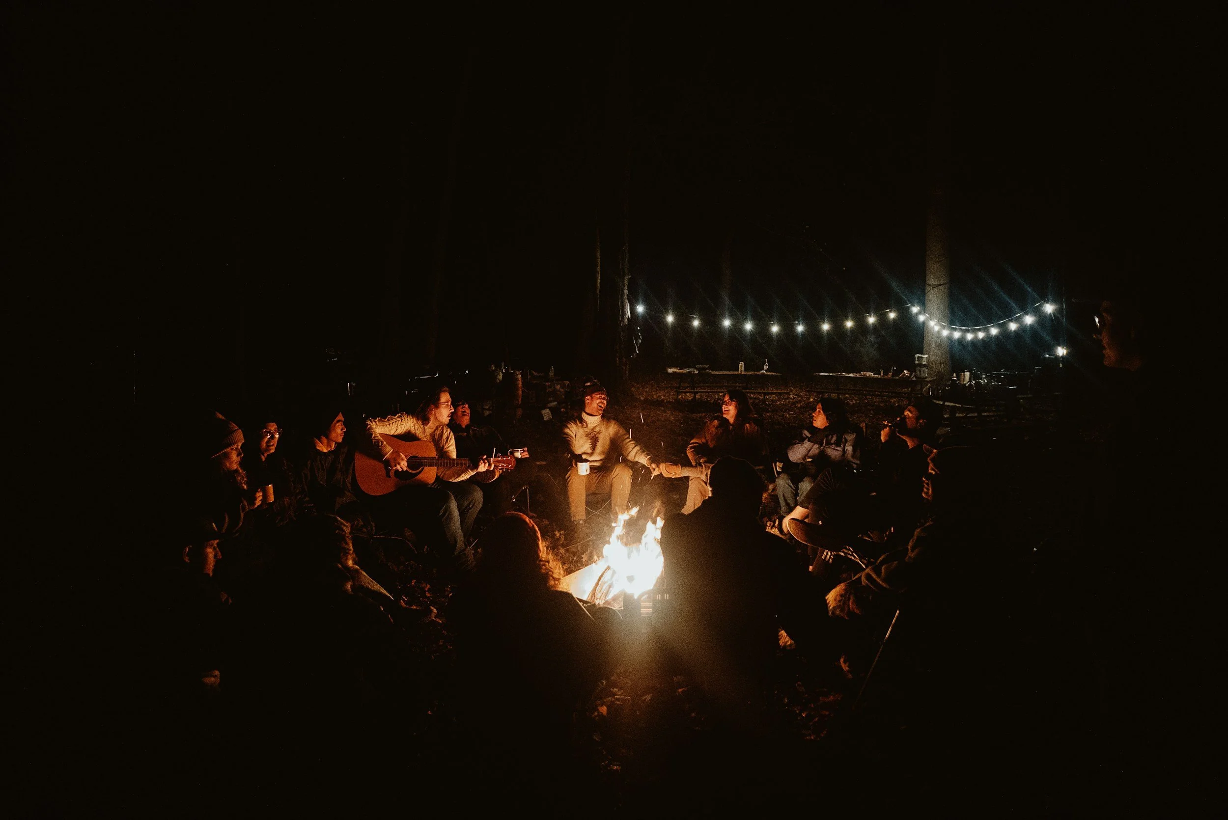 Group of people sitting around a campfire at night, some playing guitar, in an outdoor setting decorated with string lights.