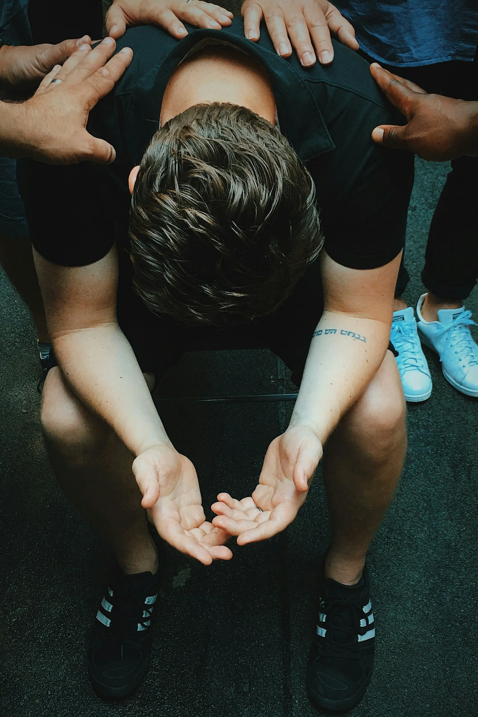A person with blonde hair is seated on the ground with their knees bent, arms extended forward, and head bowed down. Multiple hands of different skin tones are placed on the person's shoulders and head. The person is wearing a black shirt, black shorts, and black sneakers with white stripes and white soles. The background is dark ground, possibly asphalt.