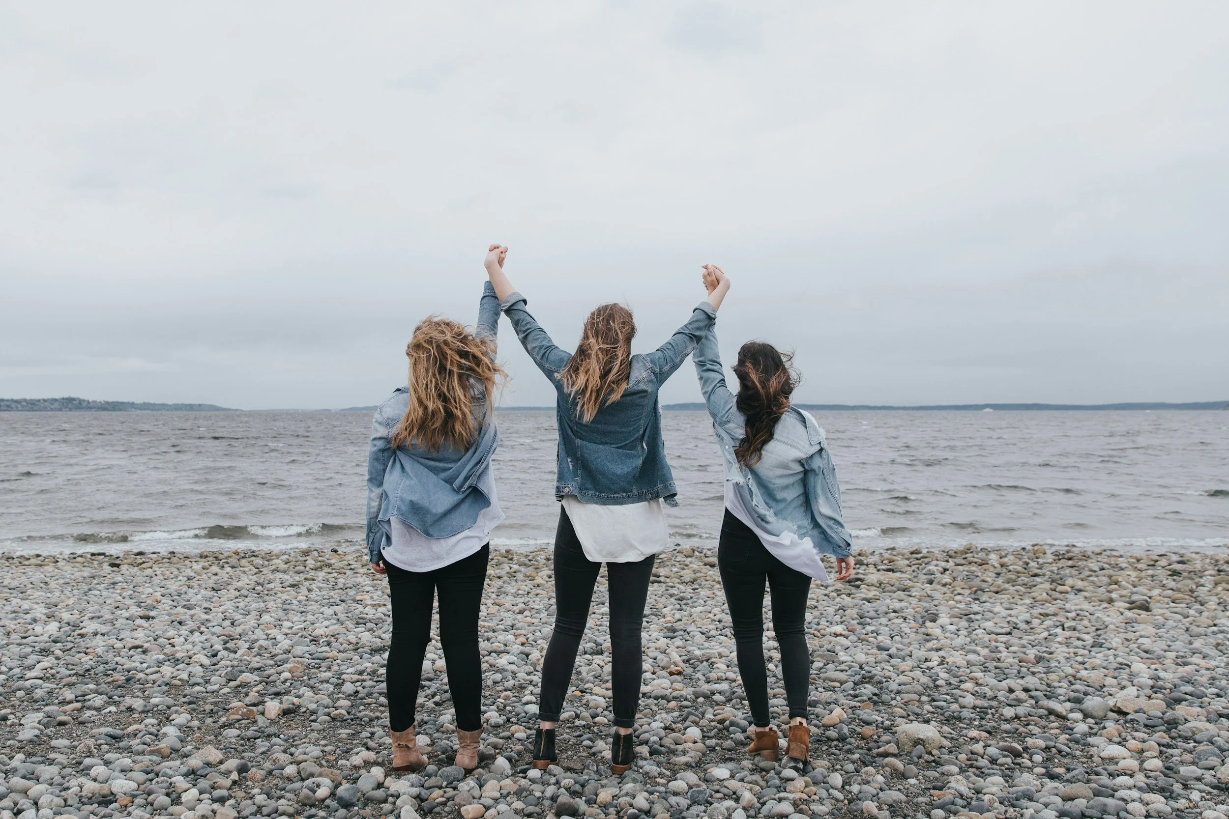 Three women wearing blue denim jackets and black pants are standing on a pebble beach, holding hands with arms raised, facing the water and cloudy sky.
