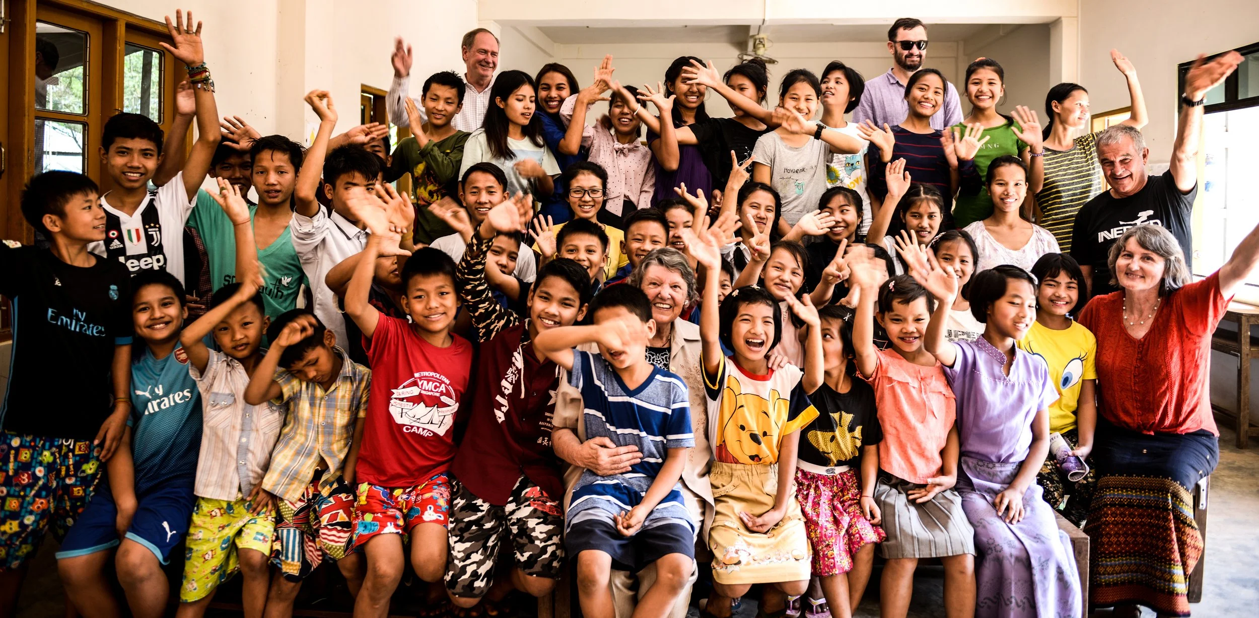 A large group of children from Mustard Seed Orphanage and a few adults posing together indoors, smiling and waving at the camera.