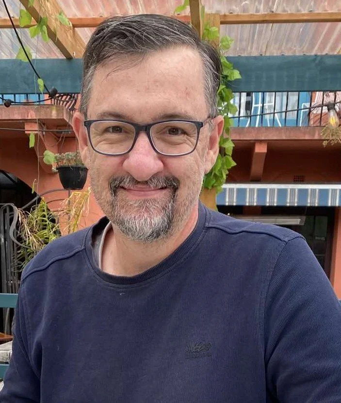 Darren Burnett, Senior Pastor, A middle-aged man with glasses, short dark hair, and a beard, smiling outdoors in front of a wooden fence with green plants and a shaded patio.