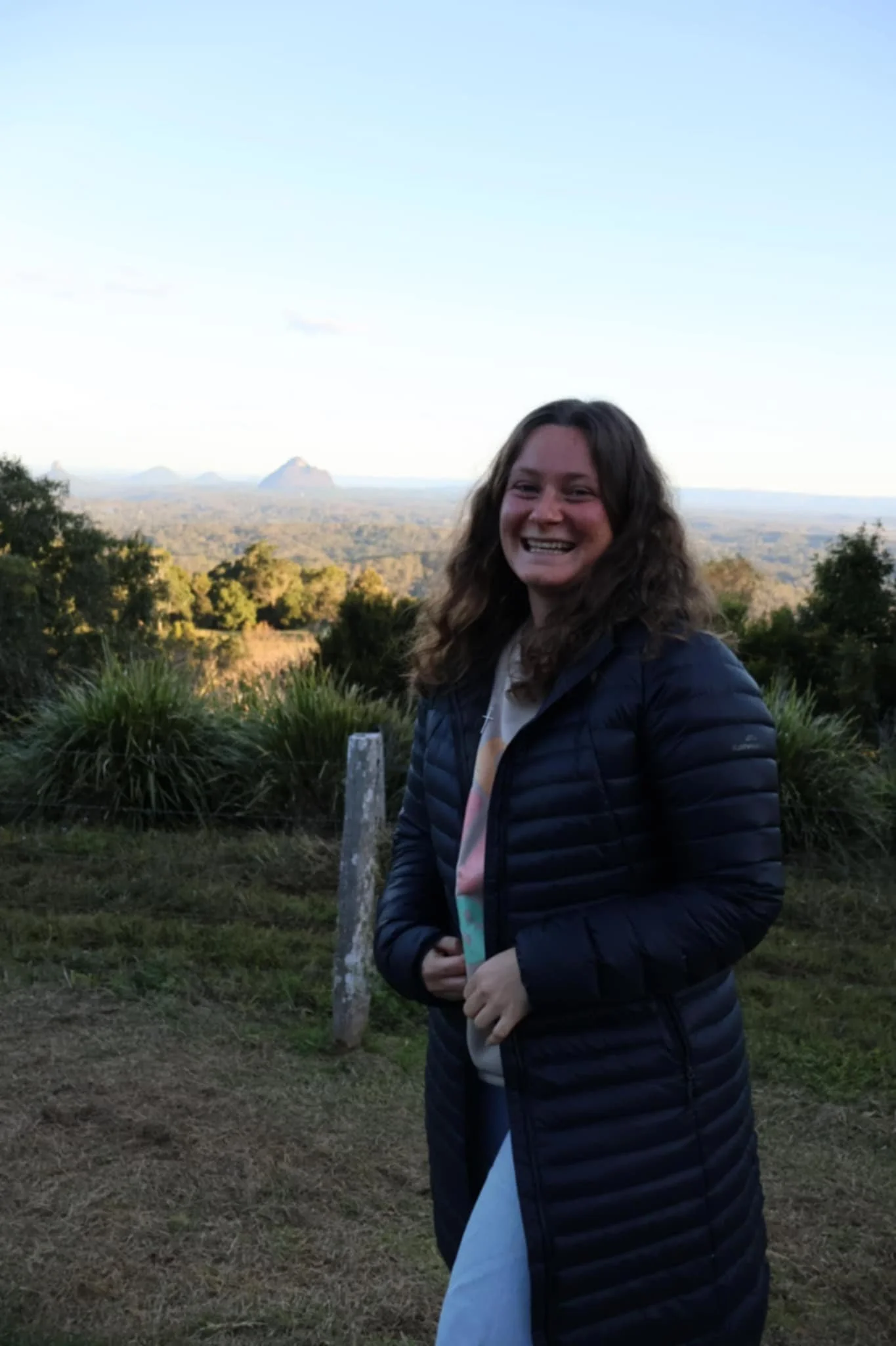 Mikeelie Ball, Youth Group Leader, A smiling woman with curly brown hair standing outdoors with a scenic landscape of trees and mountains in the background, during what appears to be late afternoon or early evening.