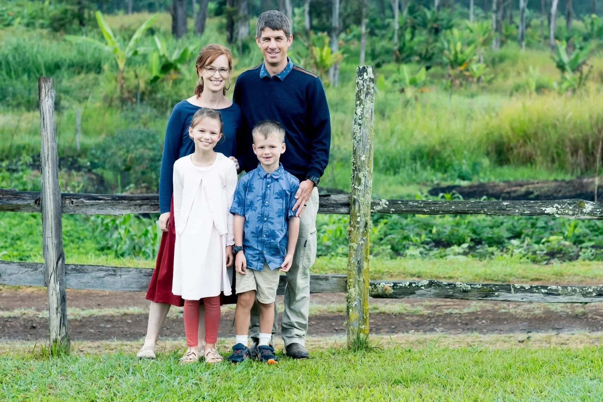 The Handasyde's, a missionary family working in PNG with Wycliffe Bible Translators and JAARS. The photo shows them standing in front of a rustic wooden fence outdoors, with lush green trees and plants in the background.