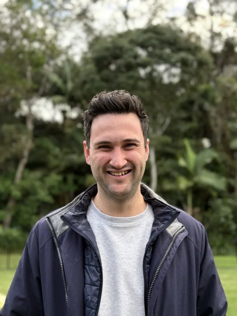 Matt Bradley, Youth Group Leader,  A smiling man outdoors in front of green trees, wearing a gray t-shirt and a navy jacket.