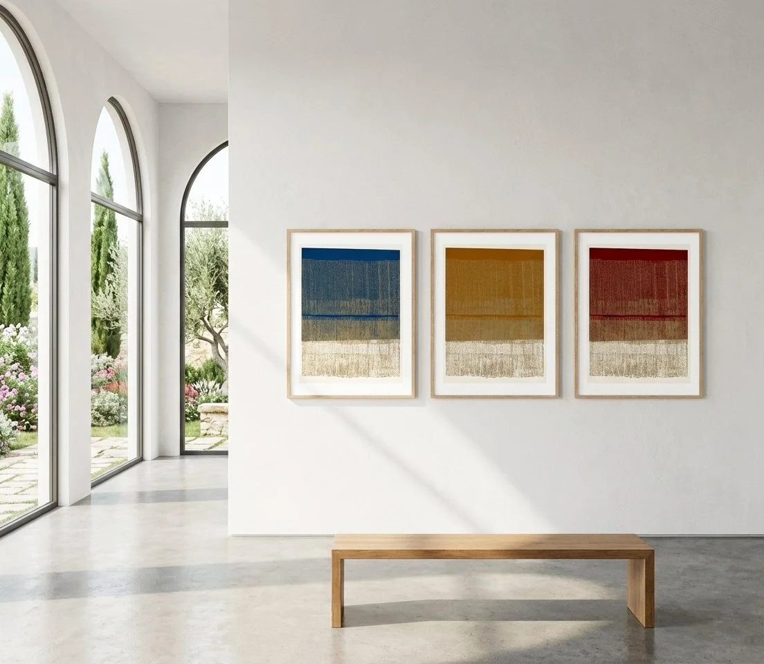 A modern art gallery with three framed abstract paintings on a white wall. The artworks feature horizontal bands of colors: blue, yellow/orange, and red. A simple wooden bench is placed below the paintings, and large arched windows reveal a landscaped garden outside.