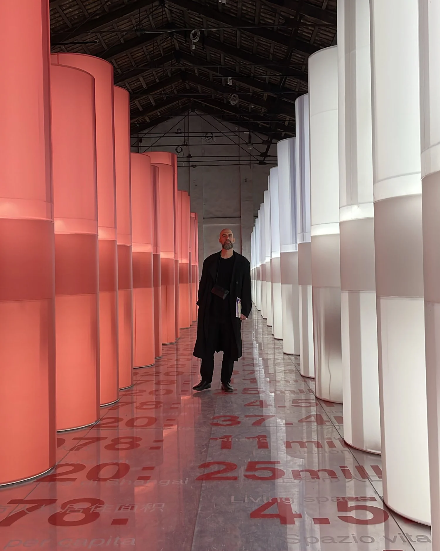 Architect Spyridon Andrikou standing in an industrial space with tall red and white light columns and a reflective floor with text.