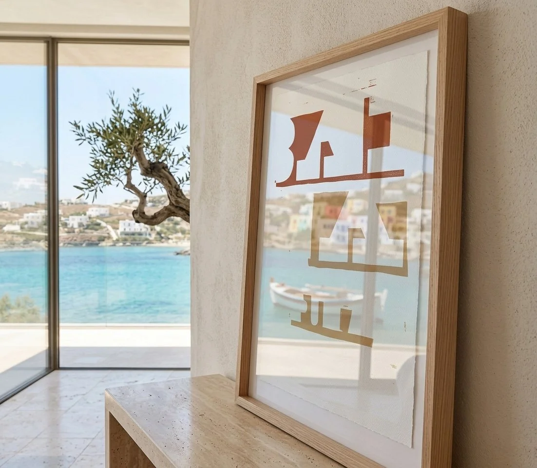 Close-up perspective of a framed nautical print on a console, showing a reflection of the blue sea and a traditional boat in the glass, with an olive tree in the background.
