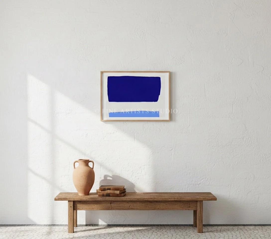 A vertical framed cobalt and sky blue abstract print styled on a minimalist shelf next to a stack of books, a small glass vase, and organic designer objects.